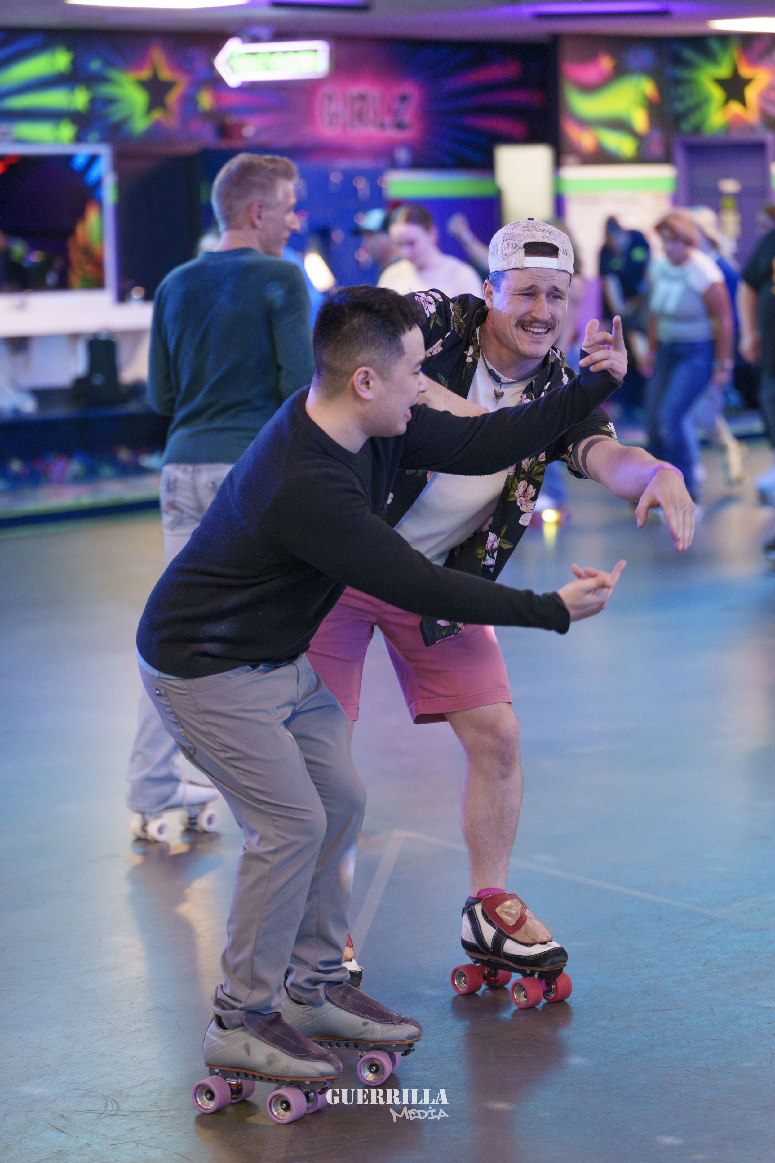 Two men roller skating and dancing together at a roller rink with colorful neon lights, while other people skate and socialize in the background.