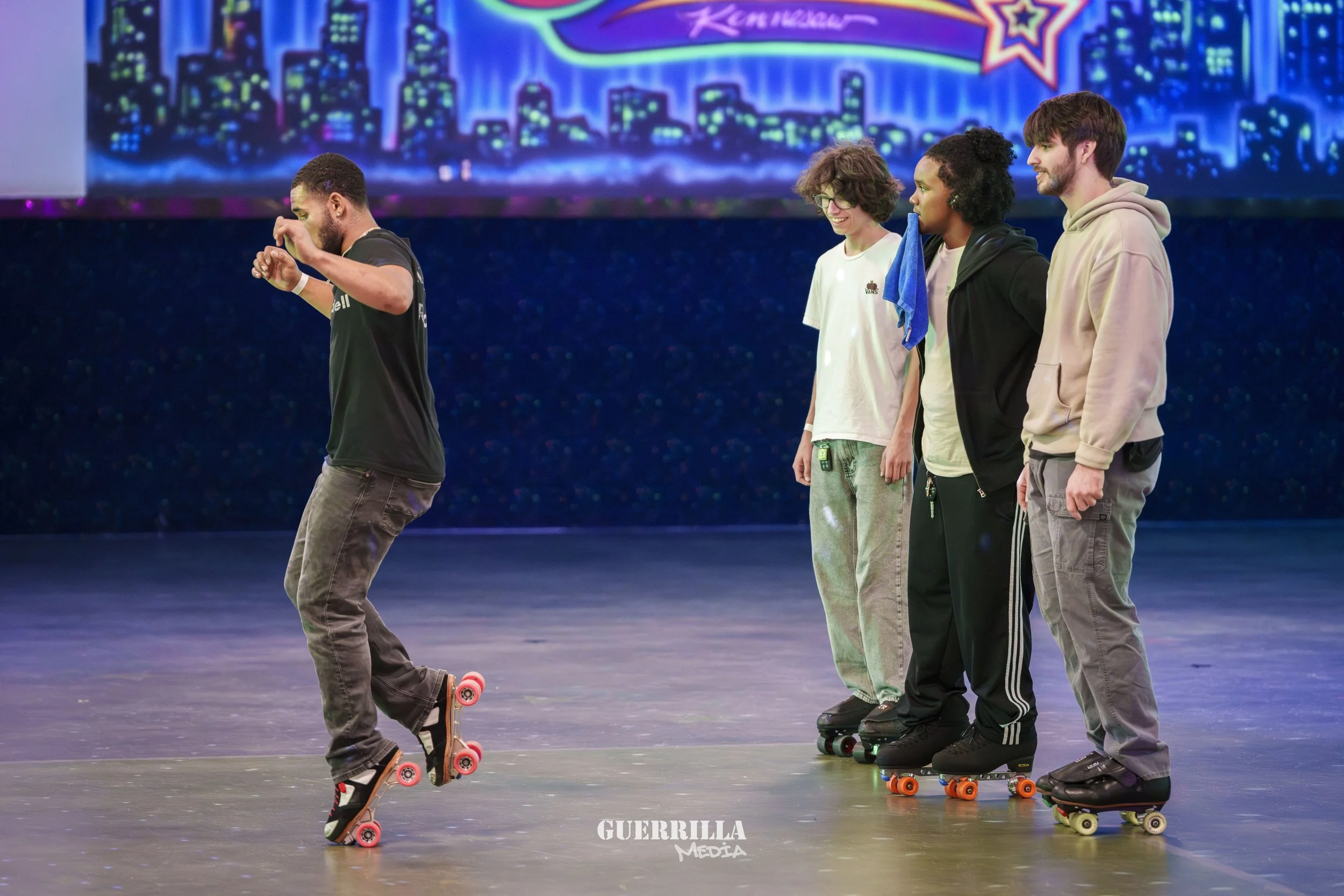 Four young people roller skating on an indoor rink, one in the front doing a trick, the other three standing in line watching. The background features a colorful cityscape mural and a bright sign that reads 'Kerrigan.'