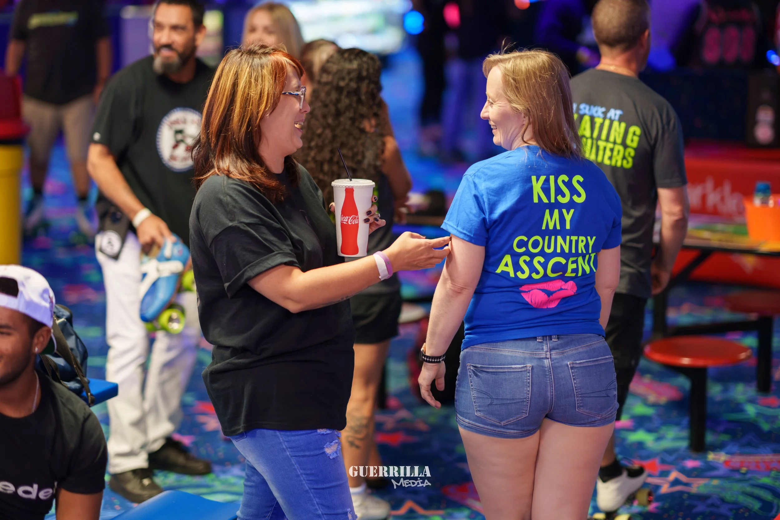 Two women laughing and talking at an indoor event, one holding a Coca-Cola cup, the other wearing a blue T-shirt with pink lips and yellow text, surrounded by other people and arcade machines.