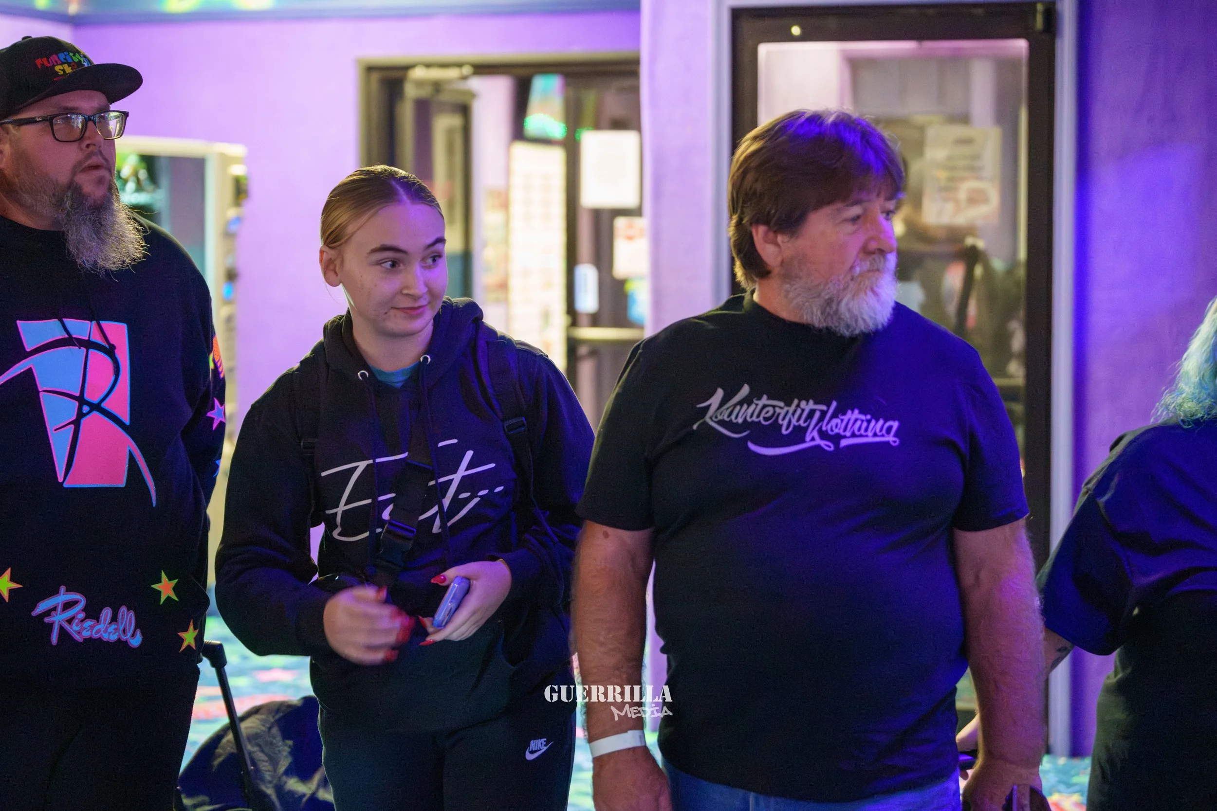 Three people standing together in a neon-lit arcade or gaming center. The person on the left is a man with a gray beard wearing glasses, a black hoodie with colorful graphics, and a black cap. The person in the middle is a young woman with blonde hai