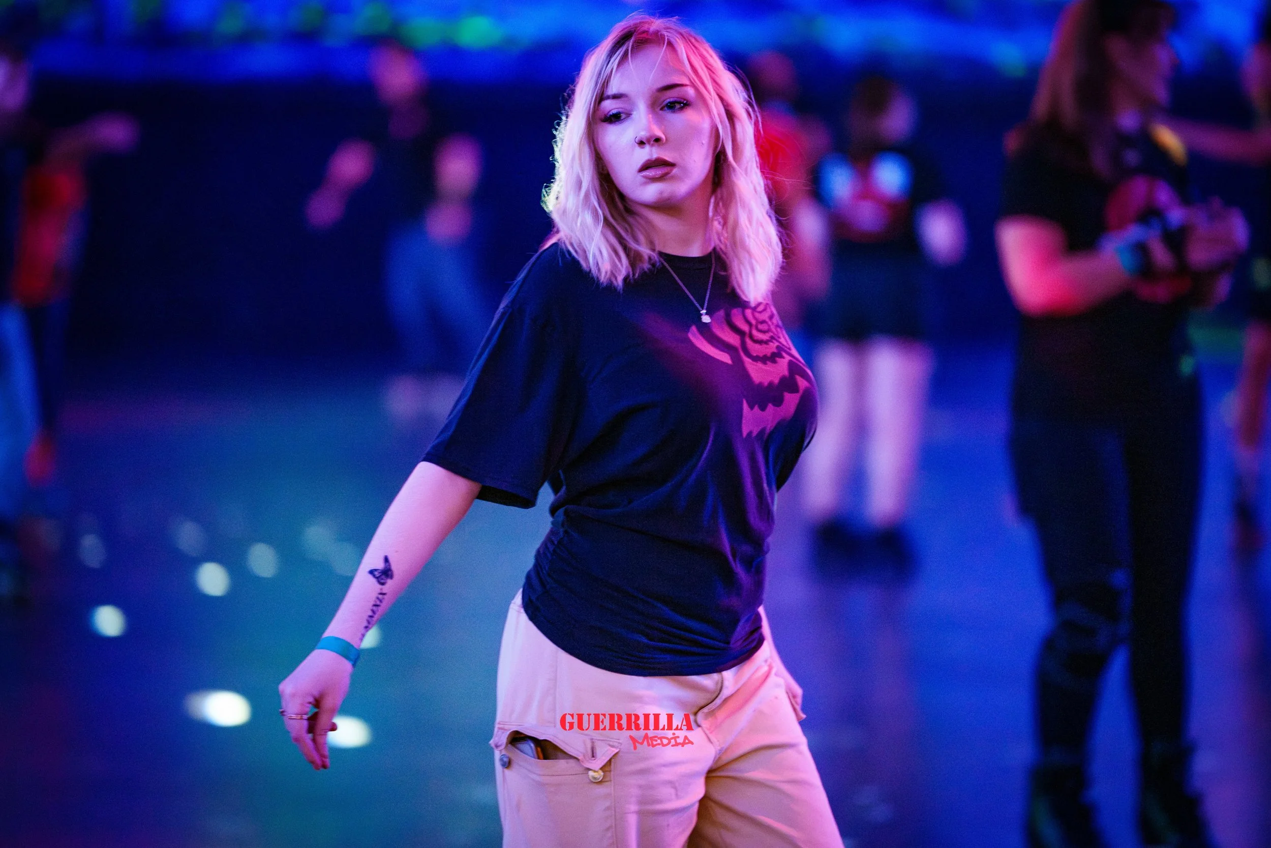A young woman with blonde, wavy hair standing in an indoor roller skating rink illuminated by colorful lights, wearing a black T-shirt with a pink graphic and beige pants with 'GUERRILLA MEDIA' written on them.