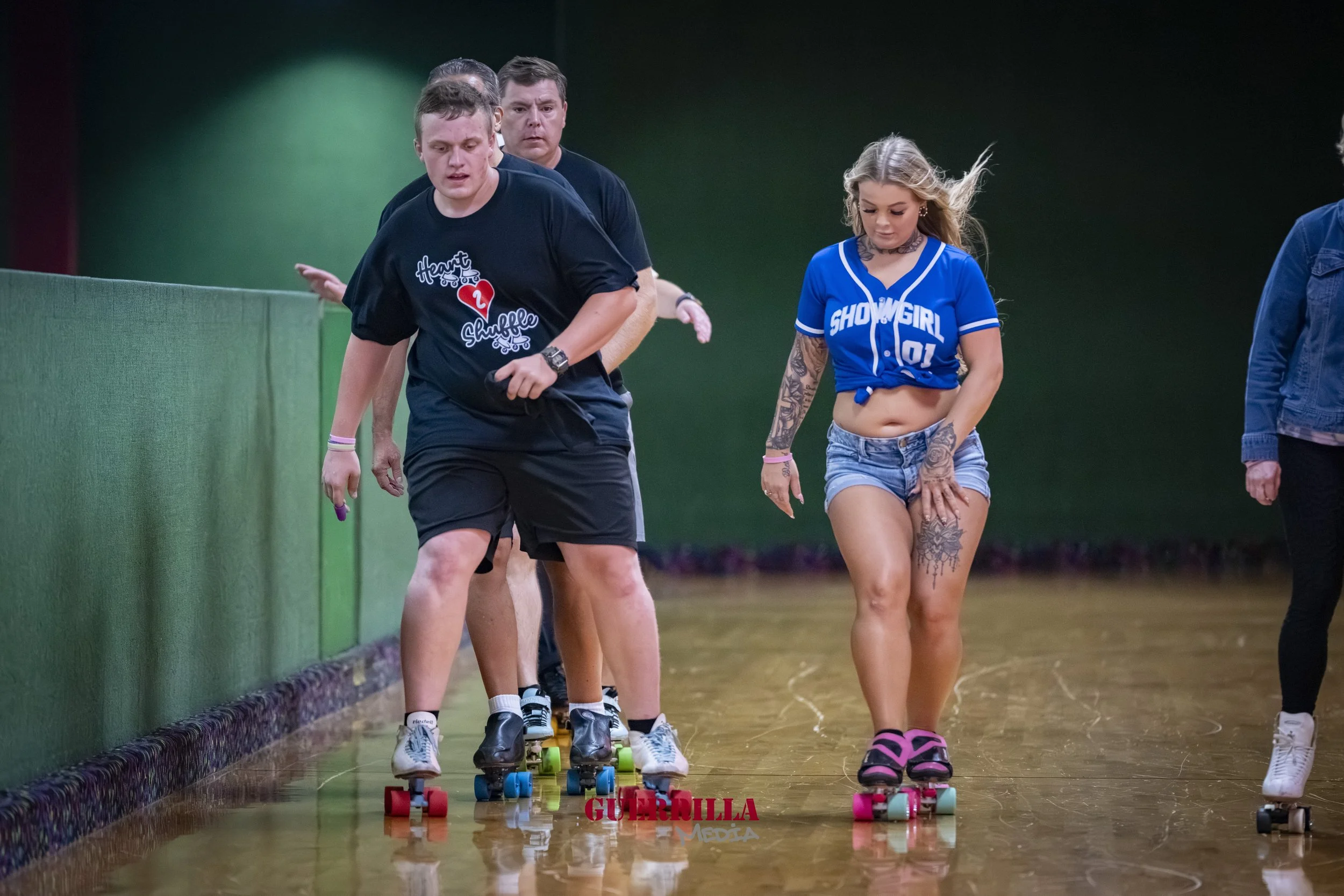 Four people roller skating on an indoor rink, with two young women and two young men, one woman has long hair and tattoos, and is wearing a blue crop top and denim shorts, the other man is wearing a black T-shirt and shorts.