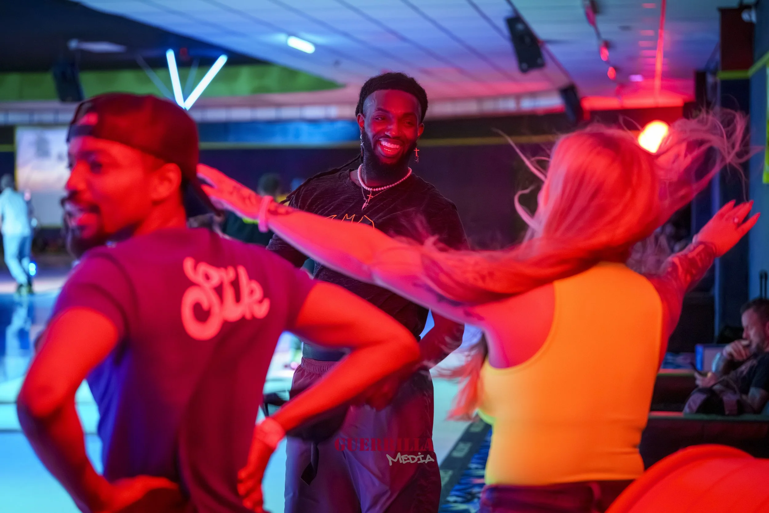 Three people dancing at a club with colorful neon lights and a DJ booth in the background. One man is smiling, wearing a black shirt and cross jewelry, while a blonde woman in a yellow top and another man in a cap dance nearby.