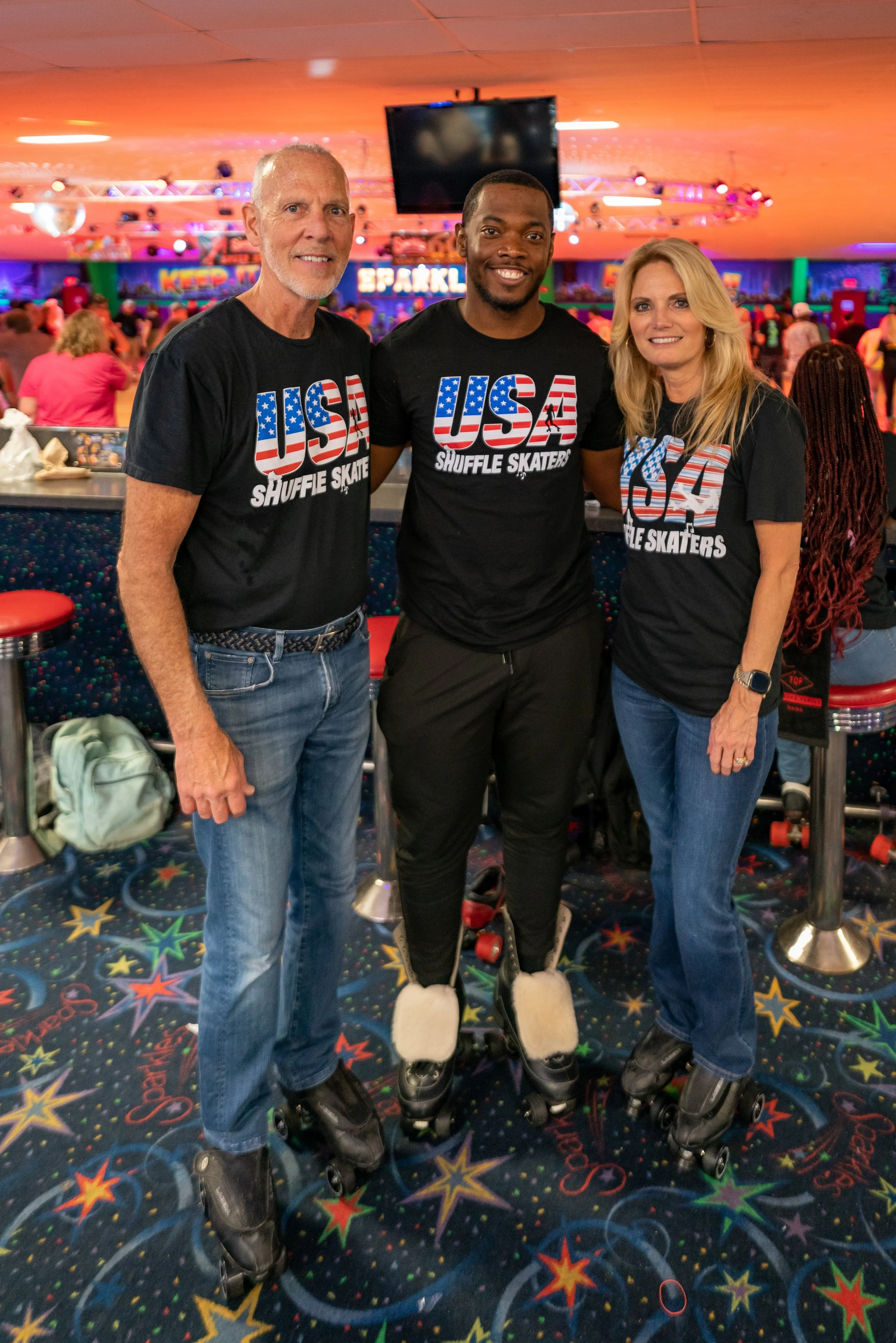 Three people wearing black USA Shuffle Skaters shirts and roller skates, standing inside a roller skating rink with a colorful, star-patterned floor and a crowd in the background.