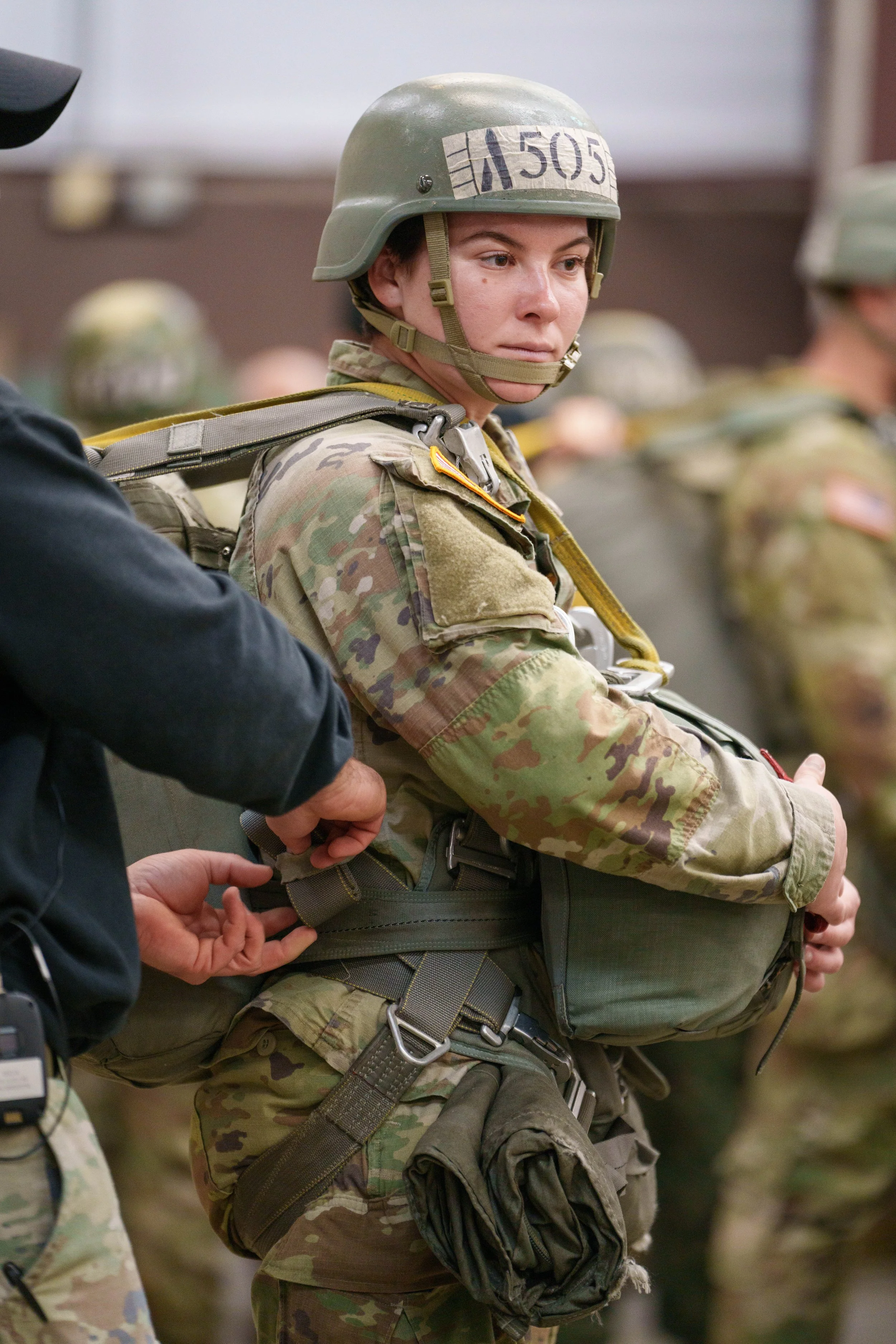 A female soldier in camouflage uniform and helmet adjusting her equipment during a military training or briefing.