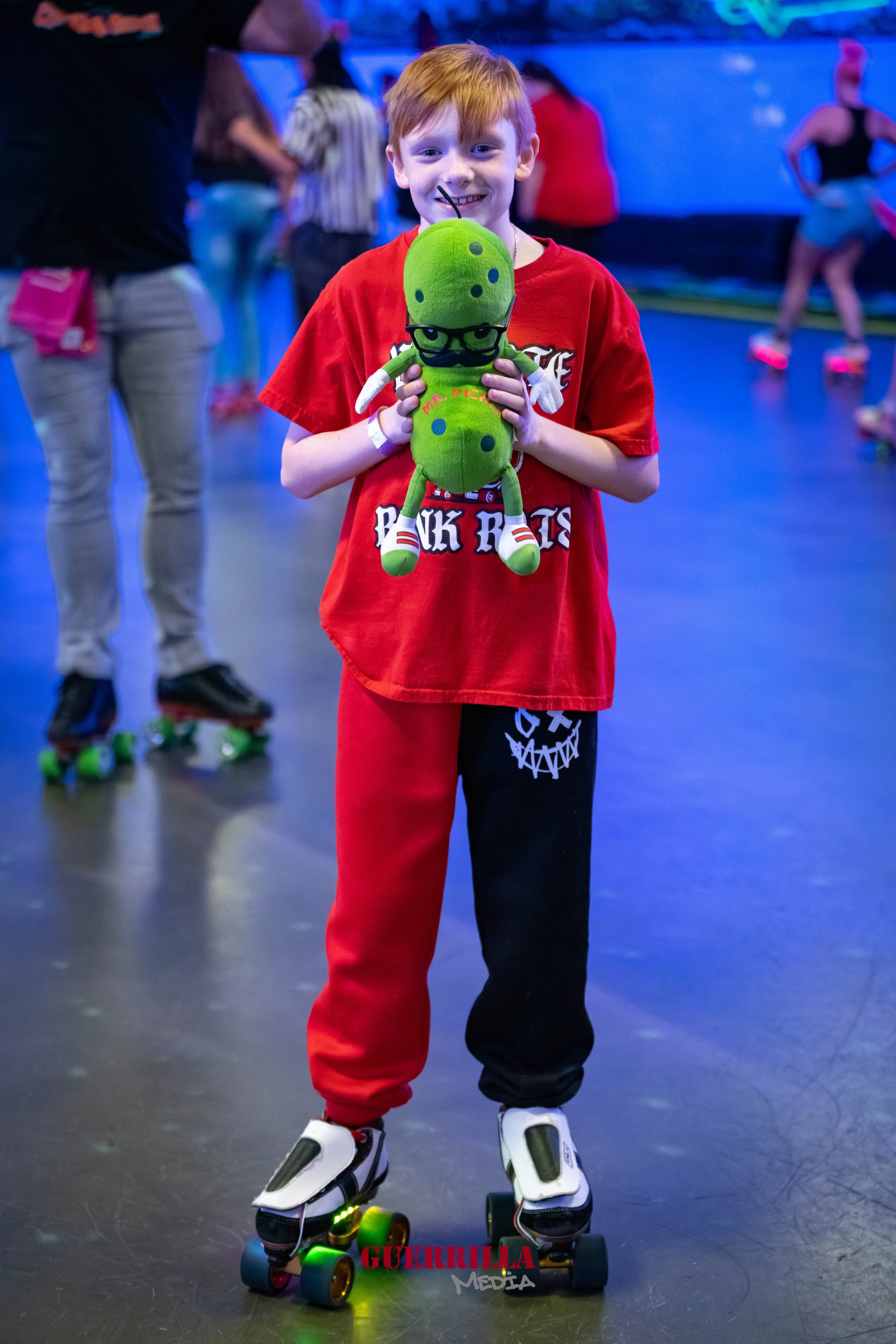 A young boy with red hair wearing a red t-shirt and black and red sweatpants is roller skating indoors. He is holding a green plush toy with glasses and black dots, smiling at the camera.