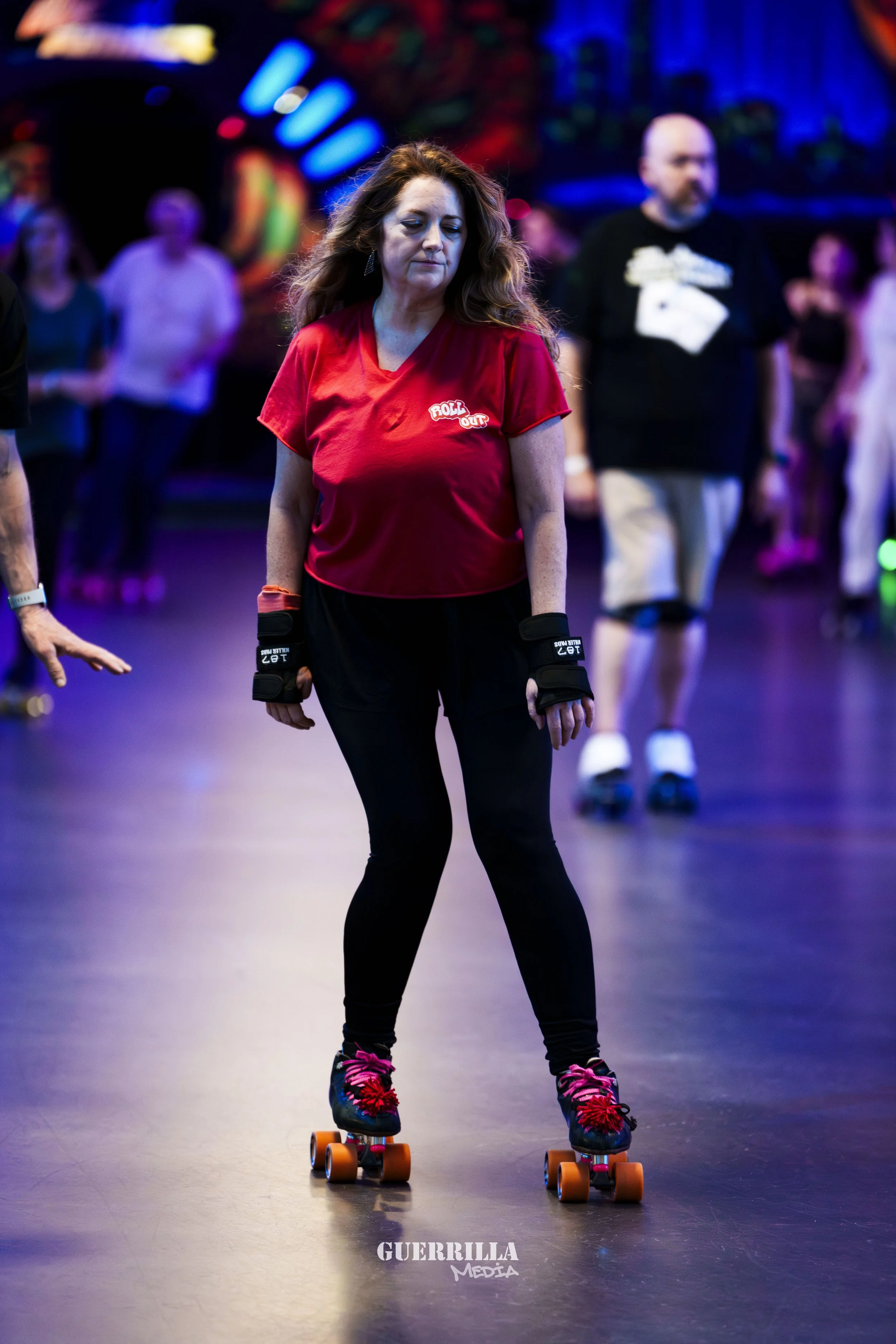 A woman roller skating with black skates and pink laces, wearing a red t-shirt with a small logo, black leggings, and protective wrist guards, in a dimly lit roller rink with colorful neon lights and other skaters in the background.
