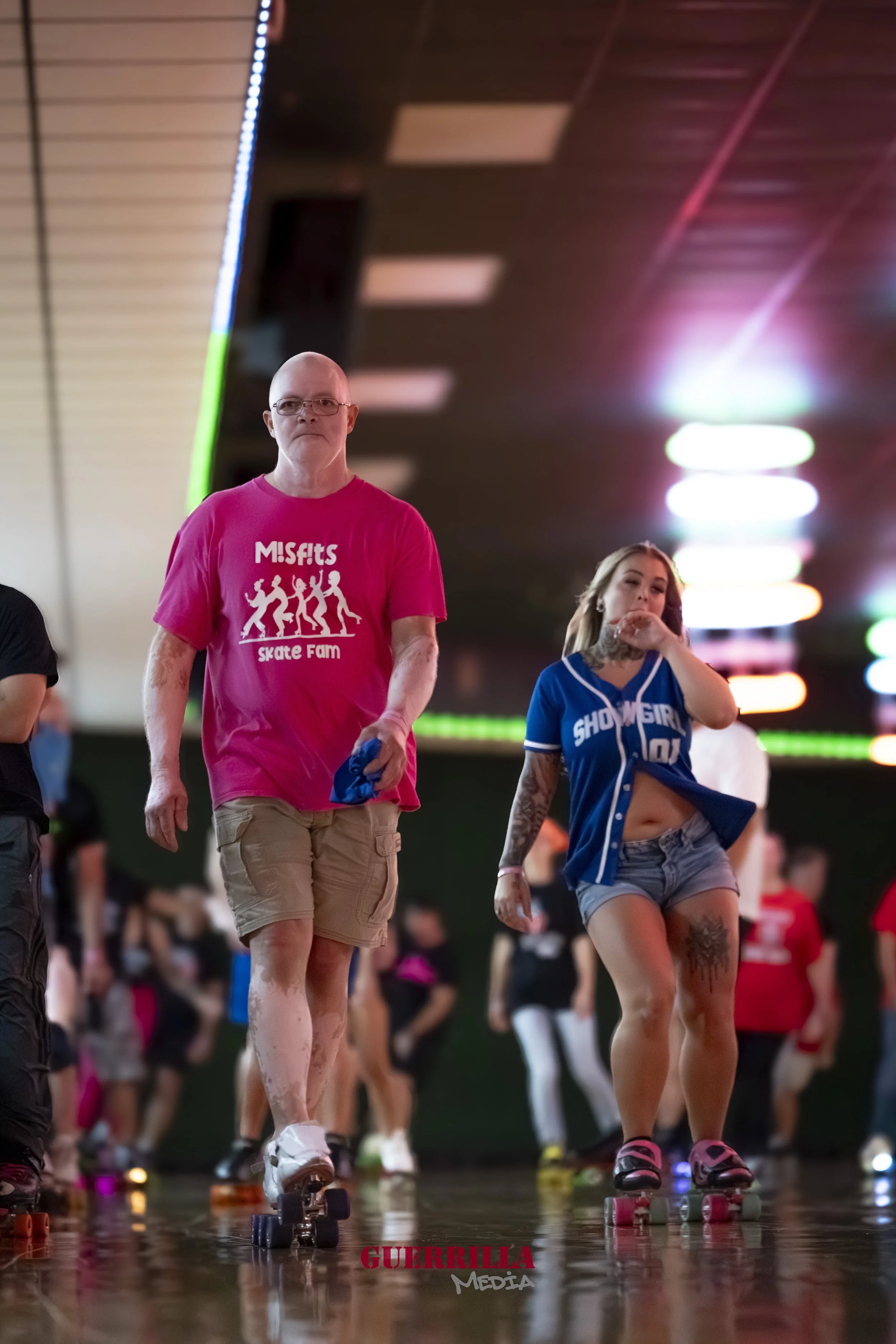 Two people roller skating in an indoor rink with colorful lights in the background. The person on the left is a man wearing glasses, a pink Misfits skate team t-shirt, and beige shorts. The person on the right is a woman with long blonde hair, wearin