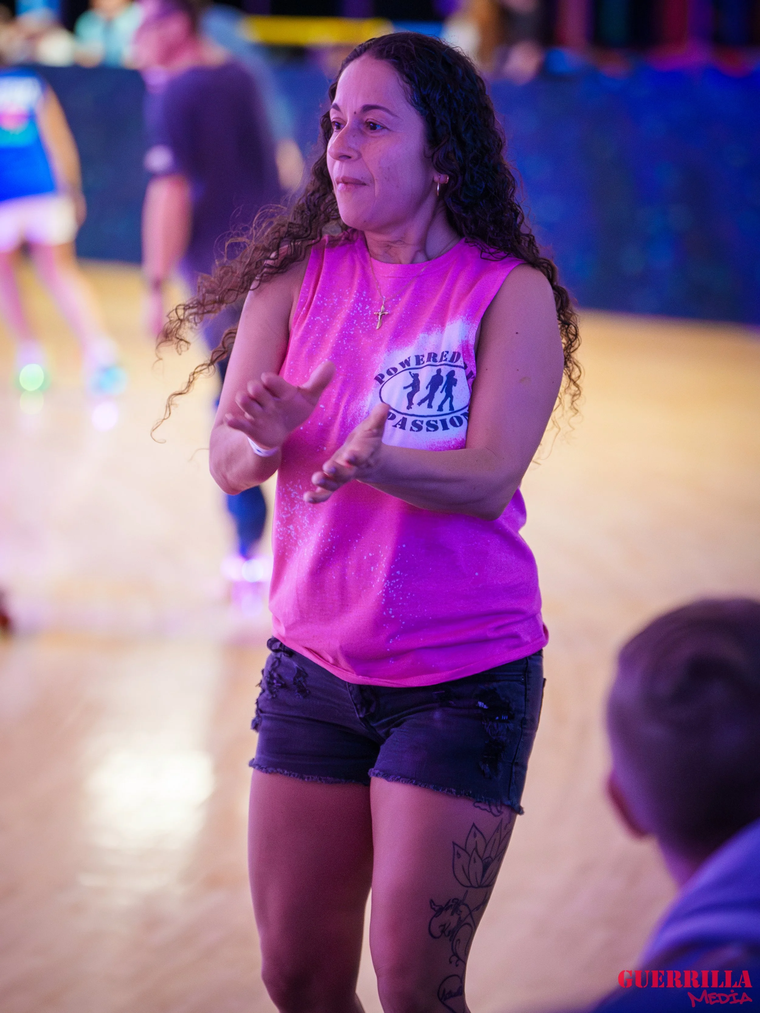 A woman with curly hair wearing a pink sleeveless shirt and black shorts, standing in a roller skating rink, clapping her hands.