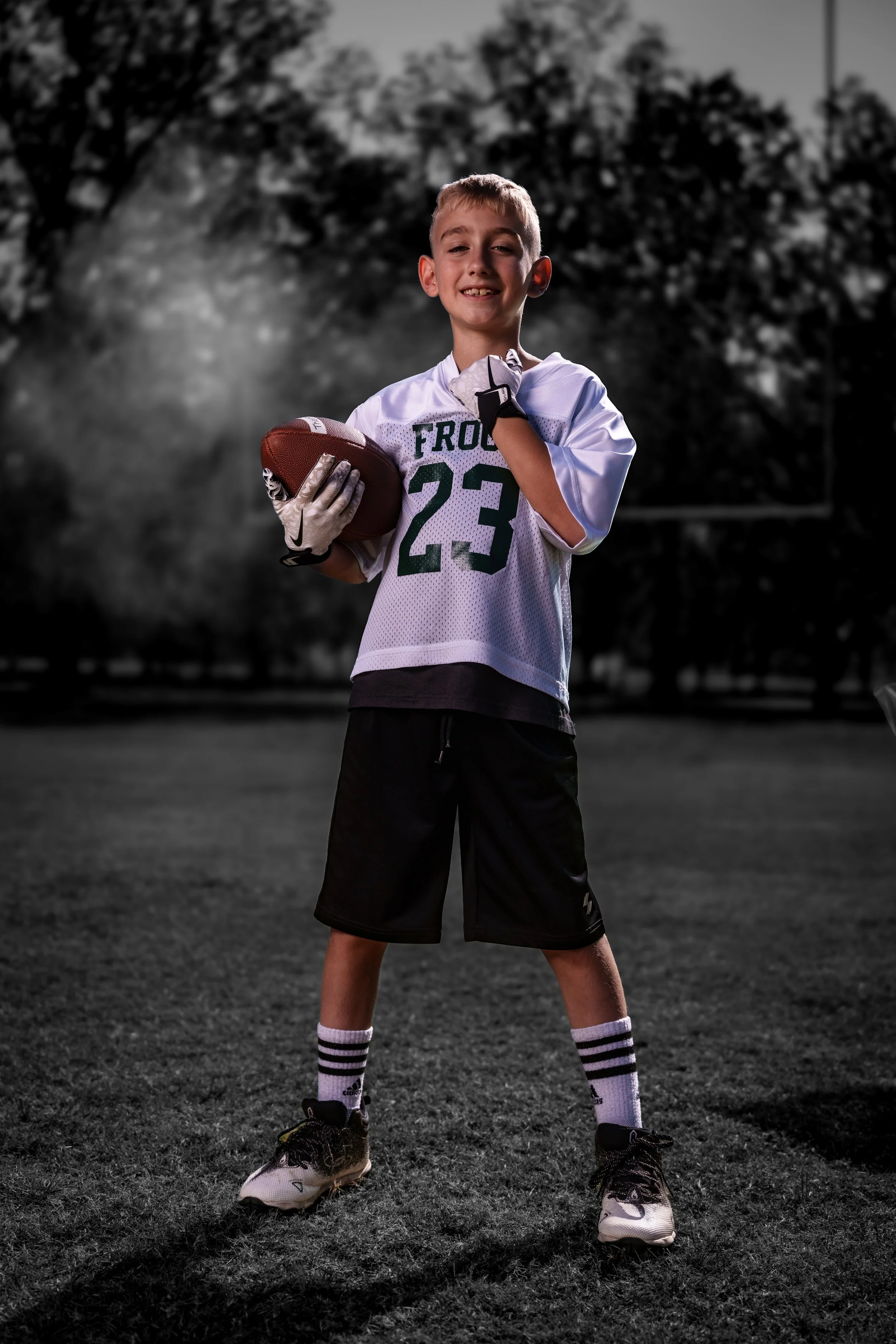 Young boy wearing a football jersey with the number 23, holding a football, standing on a grassy field with trees in the background, smiling at the camera.