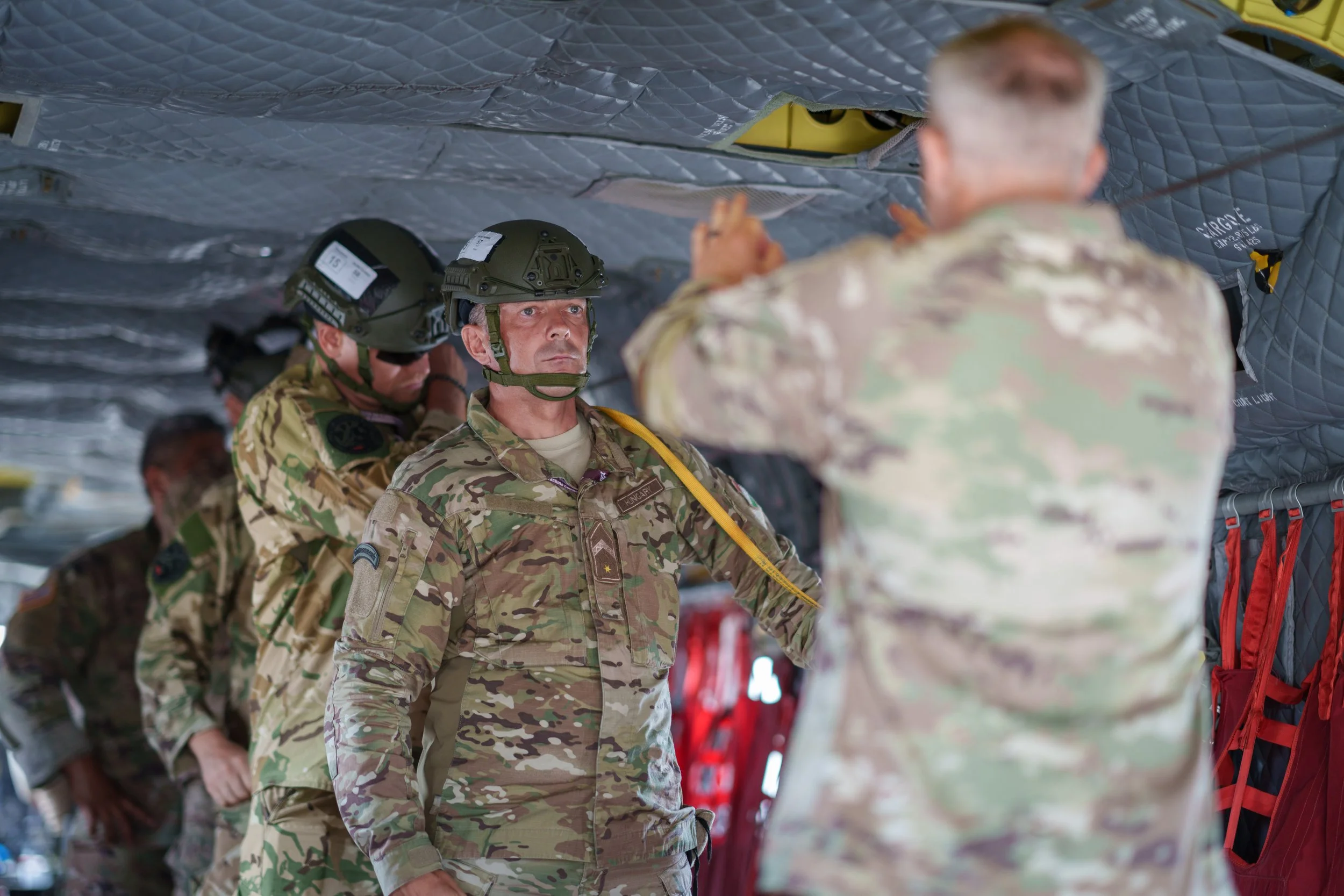 Military personnel in camouflage uniforms and helmets inside a military aircraft, listening to an officer giving instructions.