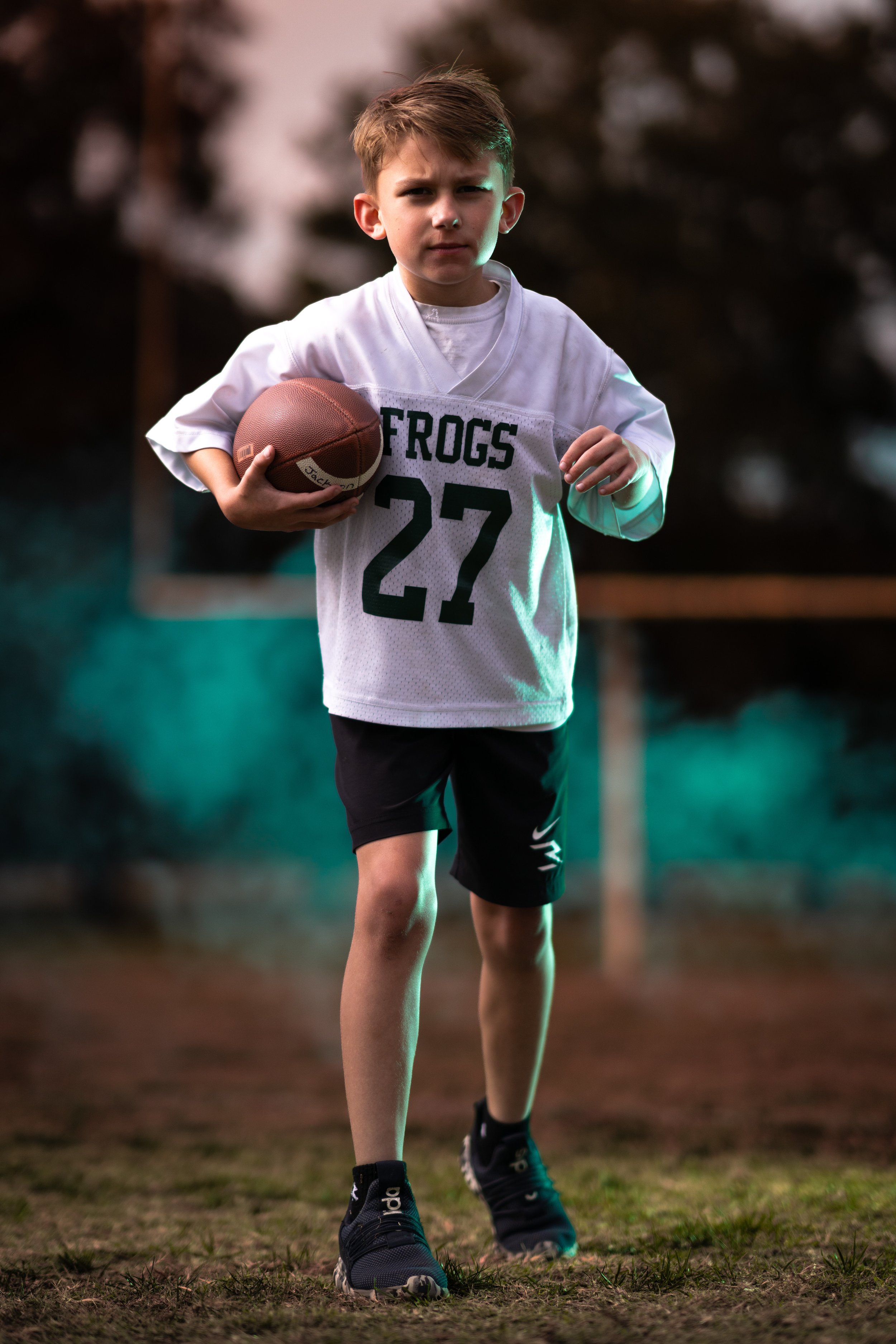 Young boy in football uniform holding a football on a field during sunset