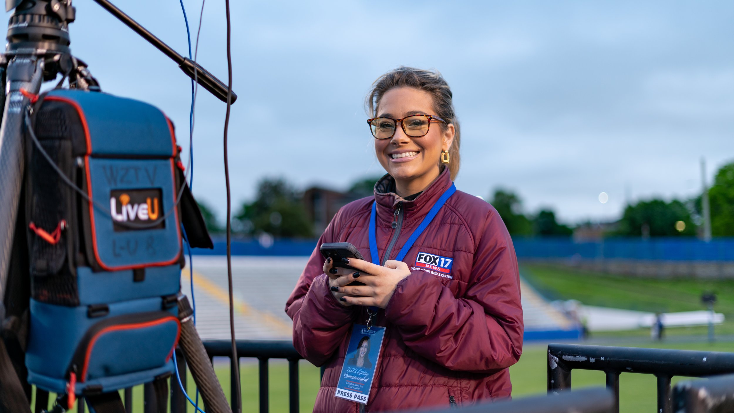 A female journalist with glasses and a maroon jacket with a FOX 17 News patch stands outdoors, smiling and holding a phone, with broadcast equipment and a press pass hanging around her neck.