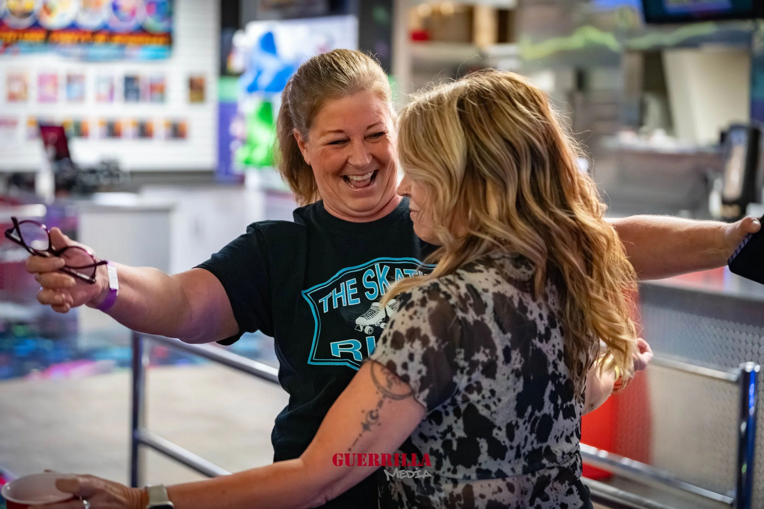 Two women embracing and smiling at each other inside a store, with one wearing glasses and a black t-shirt, and the other in a patterned shirt, with shelves of merchandise in the background.