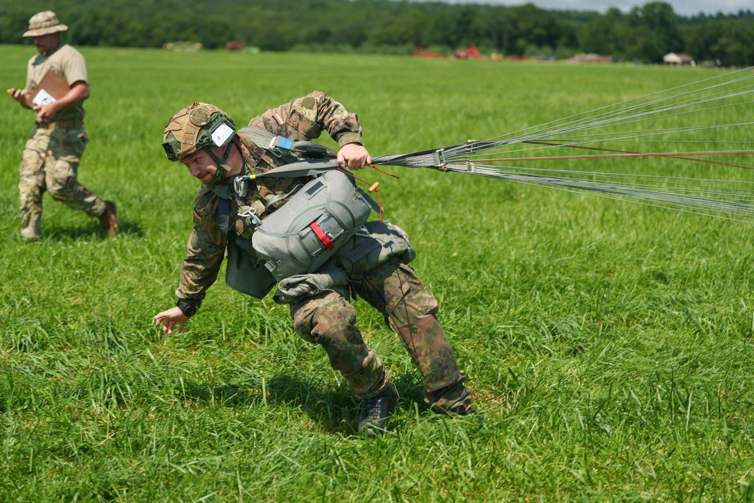 A soldier wearing camouflage uniform and gear, falling onto grass, as other personnel observe in the background, during a daytime outdoor activity.