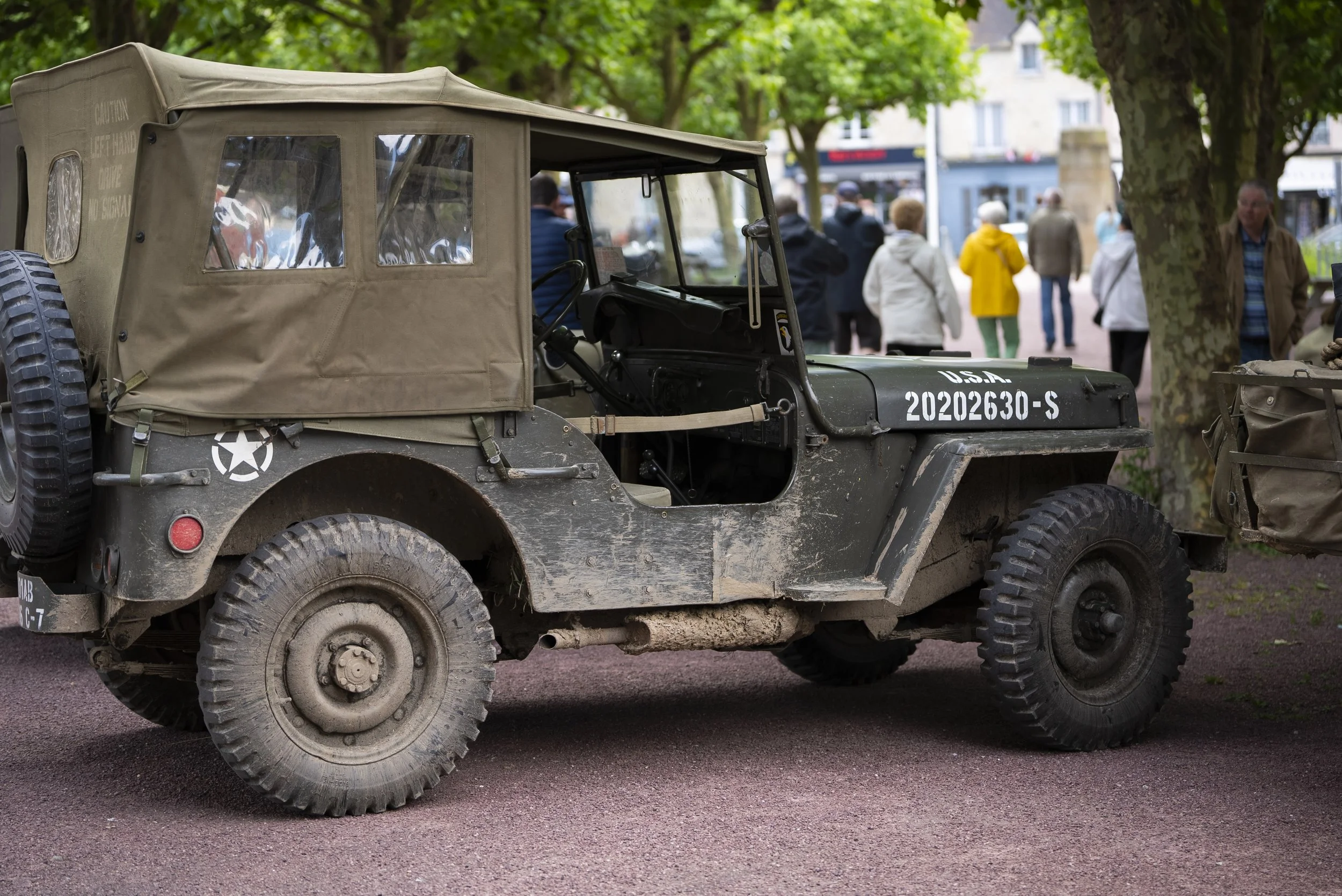 A vintage military Jeep with a tan canvas top, parked on a street with people walking and trees in the background.