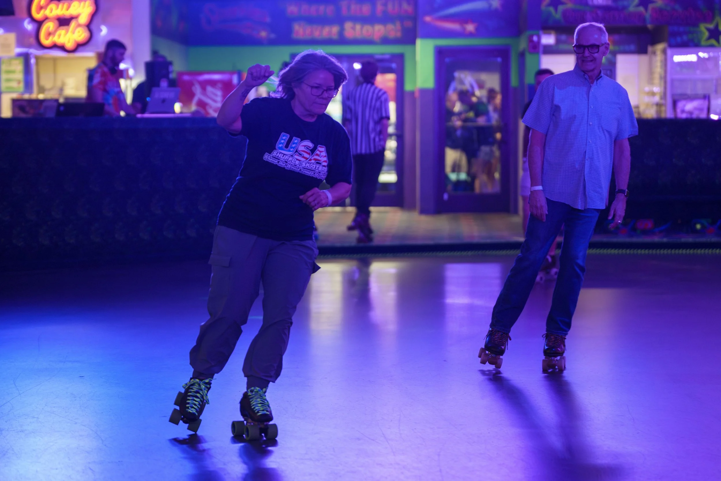 Two elderly people roller skating in an indoor rink with colorful neon lights, a woman wearing a black USA t-shirt and cargo pants, and a man in a light blue short-sleeve shirt and jeans. There are other people and arcade games in the background.