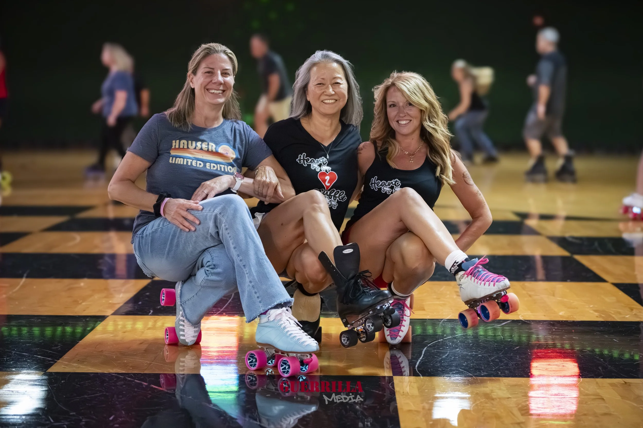 Three women sitting on roller skates on an indoor roller skating rink, smiling for the photo, with others roller skating in the background.