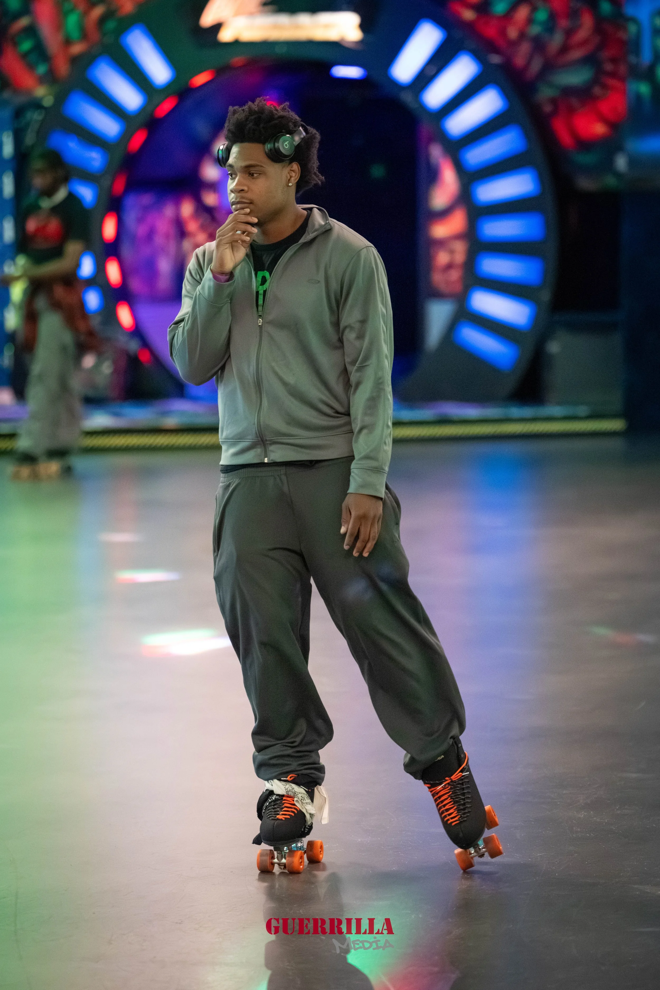 A young man skating on roller skates in an indoor entertainment center, wearing headphones, a gray zip-up jacket, black pants, and looking contemplative with his hand on his chin. The background features colorful lights and a tunnel-like decoration.