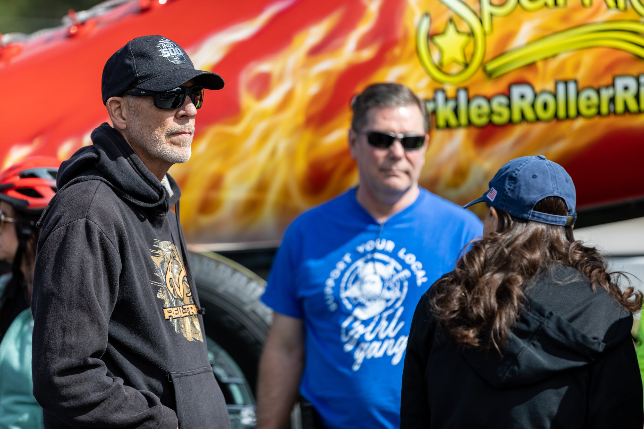 Three people standing outdoors in conversation, with an out-of-focus carnival ride or attraction in the background.