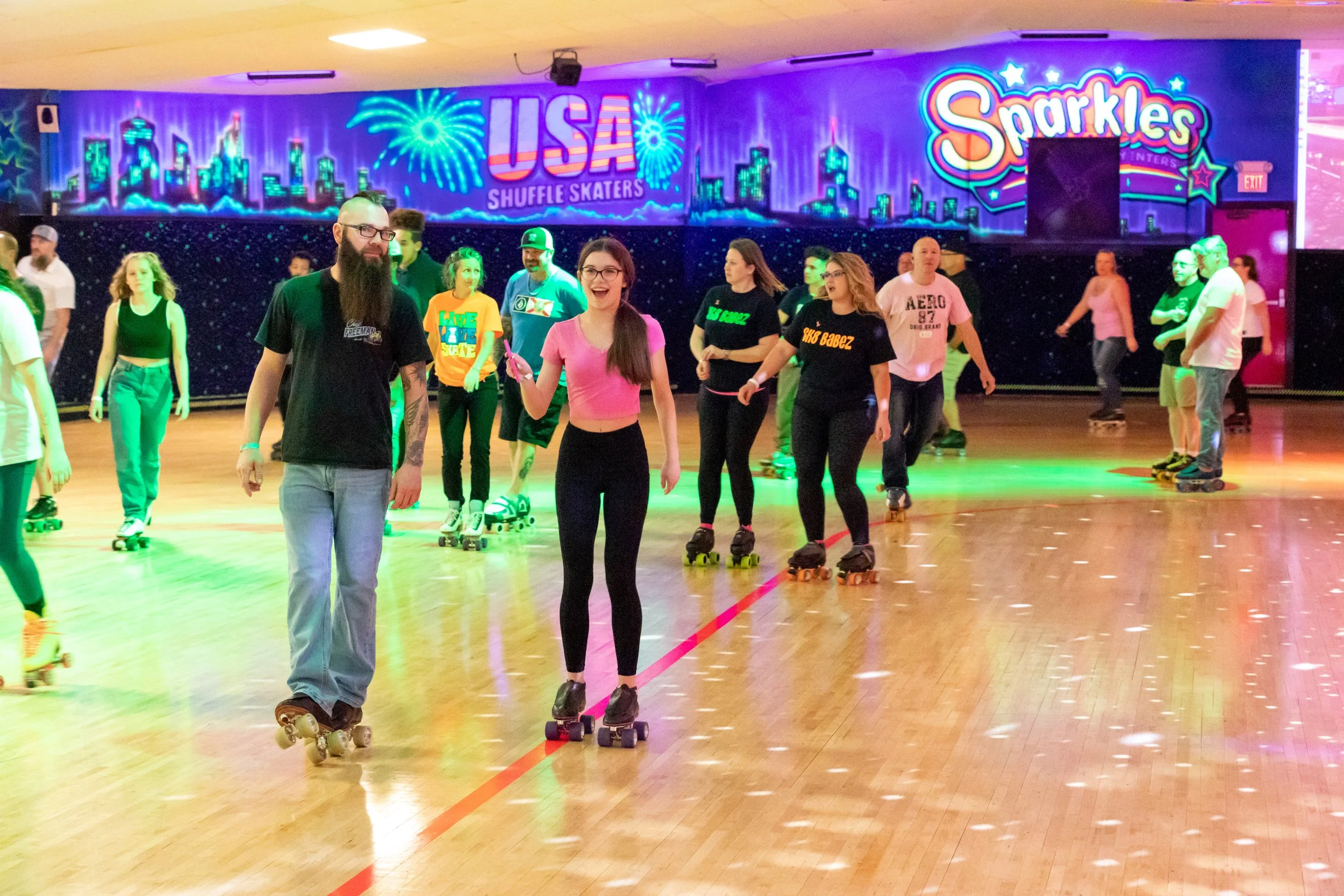 People roller skating inside an indoor rink with colorful neon signs and a cityscape mural in the background. Some skaters are smiling and enjoying the activity.