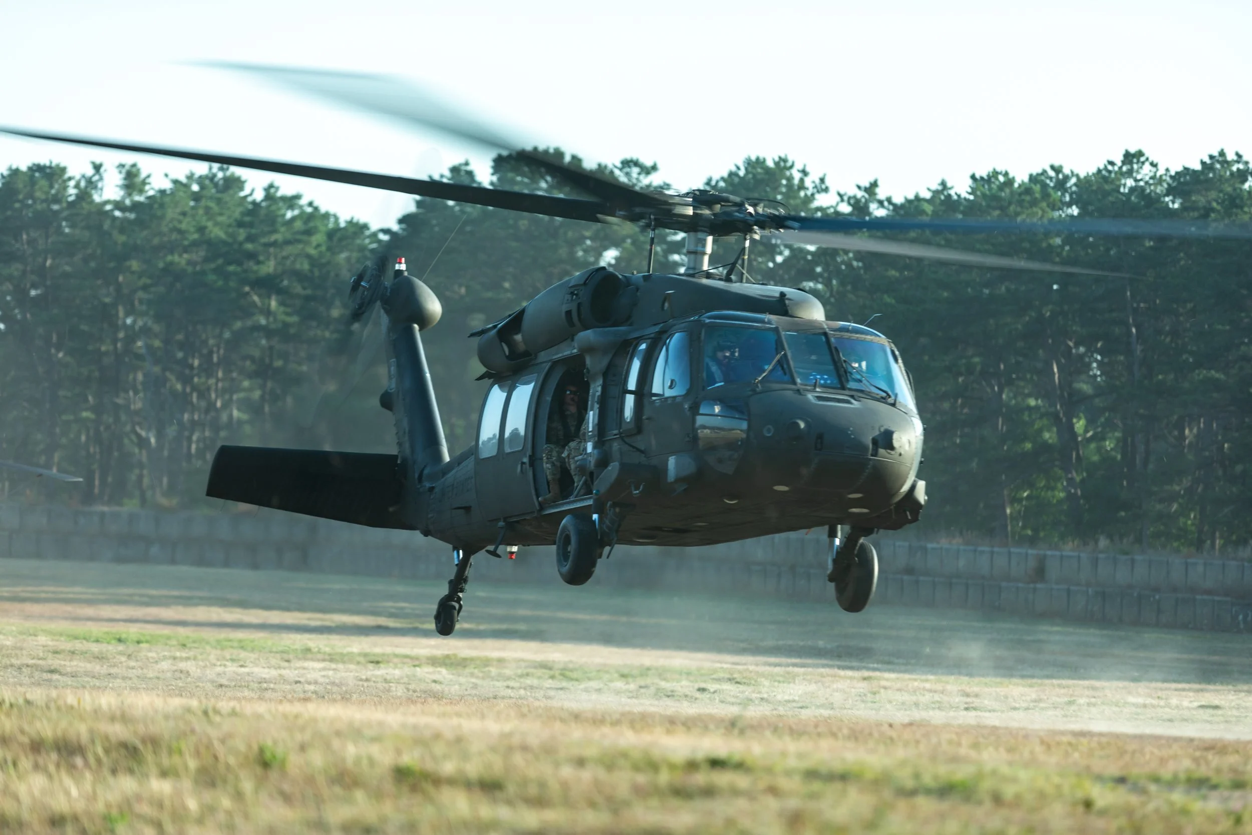 A military helicopter in mid-flight over a grassy field with trees in the background.