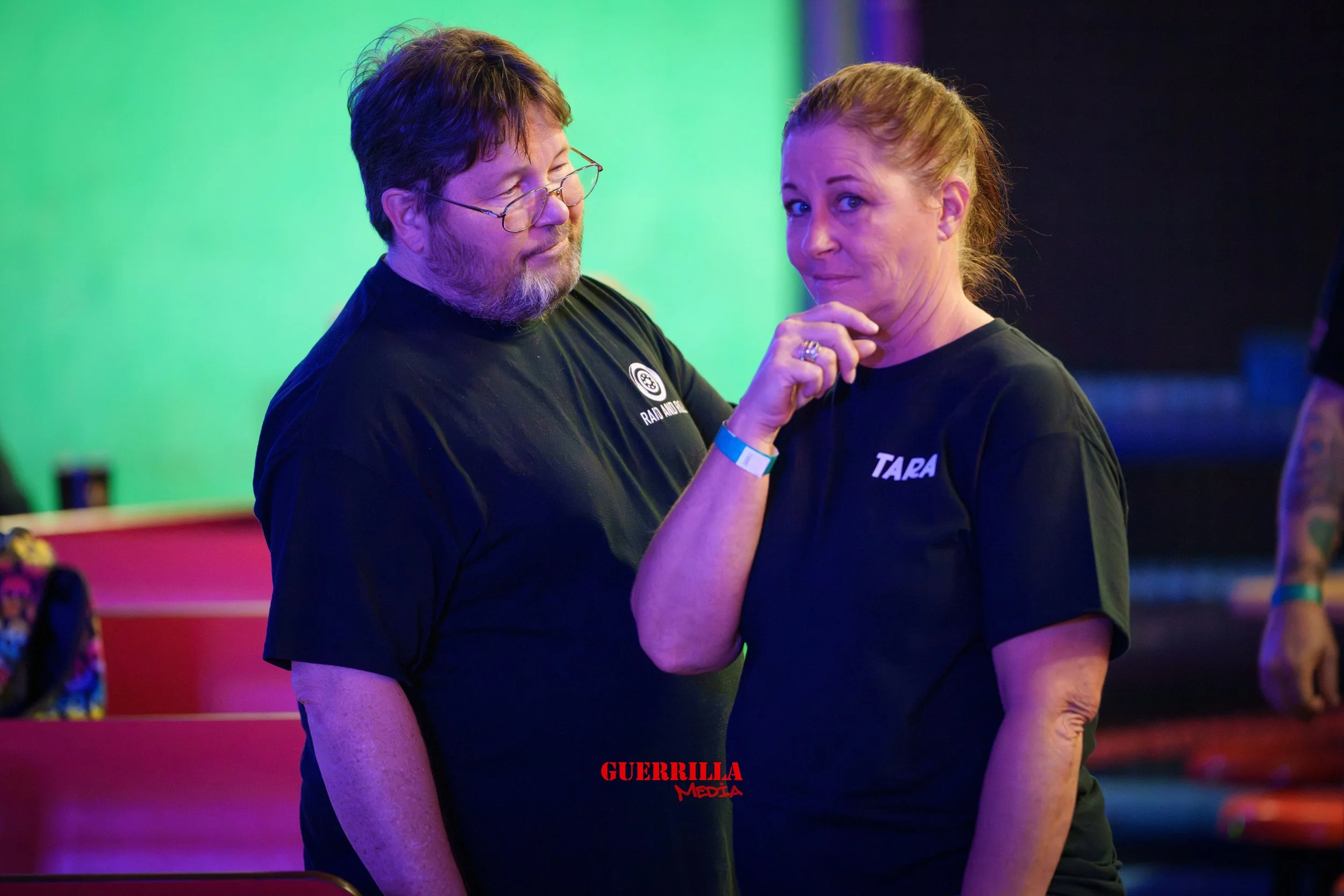 A man with glasses and a beard in a black T-shirt looking at a woman in a black T-shirt with 'TARA' printed on it, touching her chin, in a colorful indoor setting.
