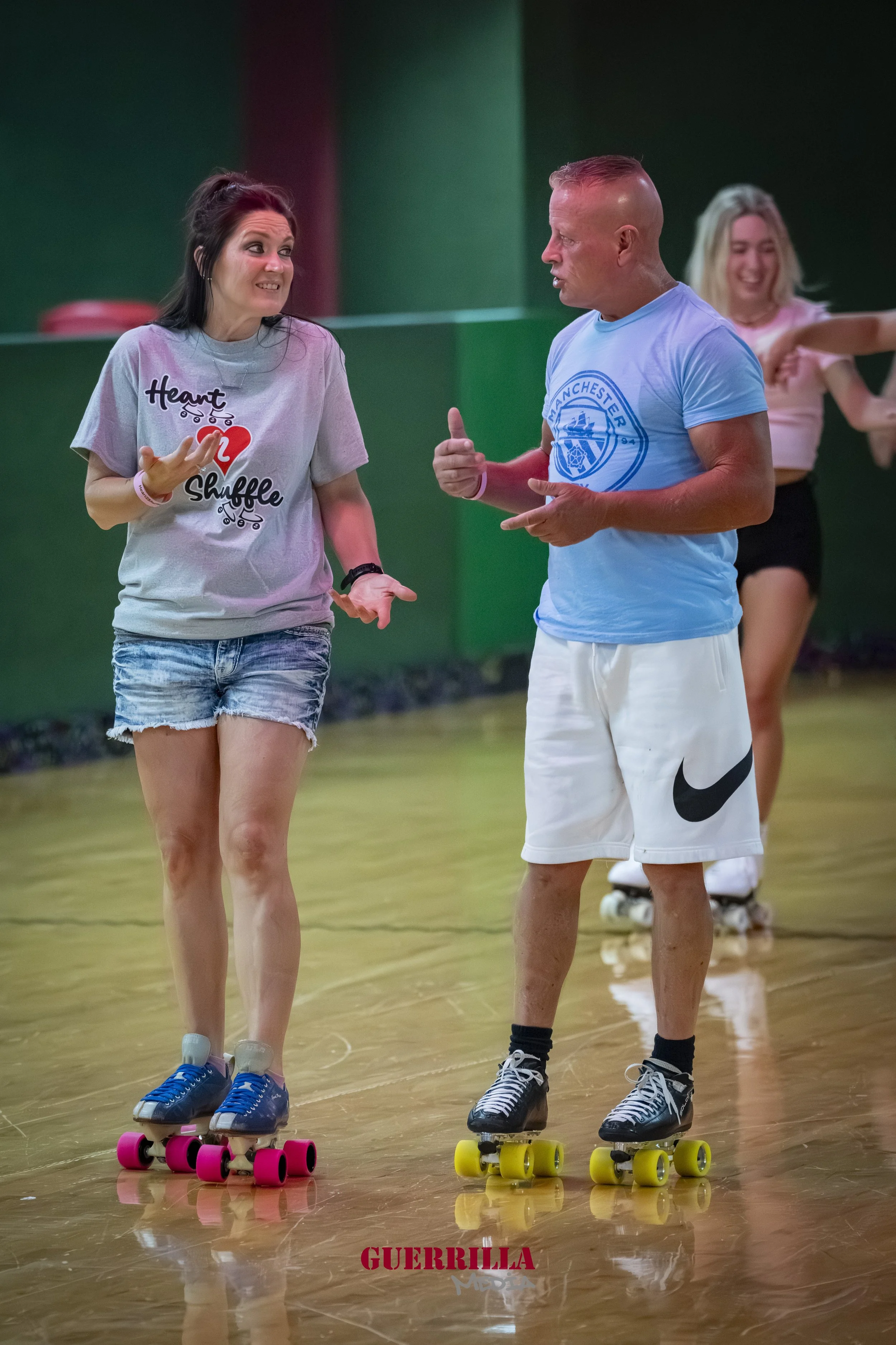 Two women and one man roller skating indoors, engaging in conversation, with a woman in the background skating and smiling.