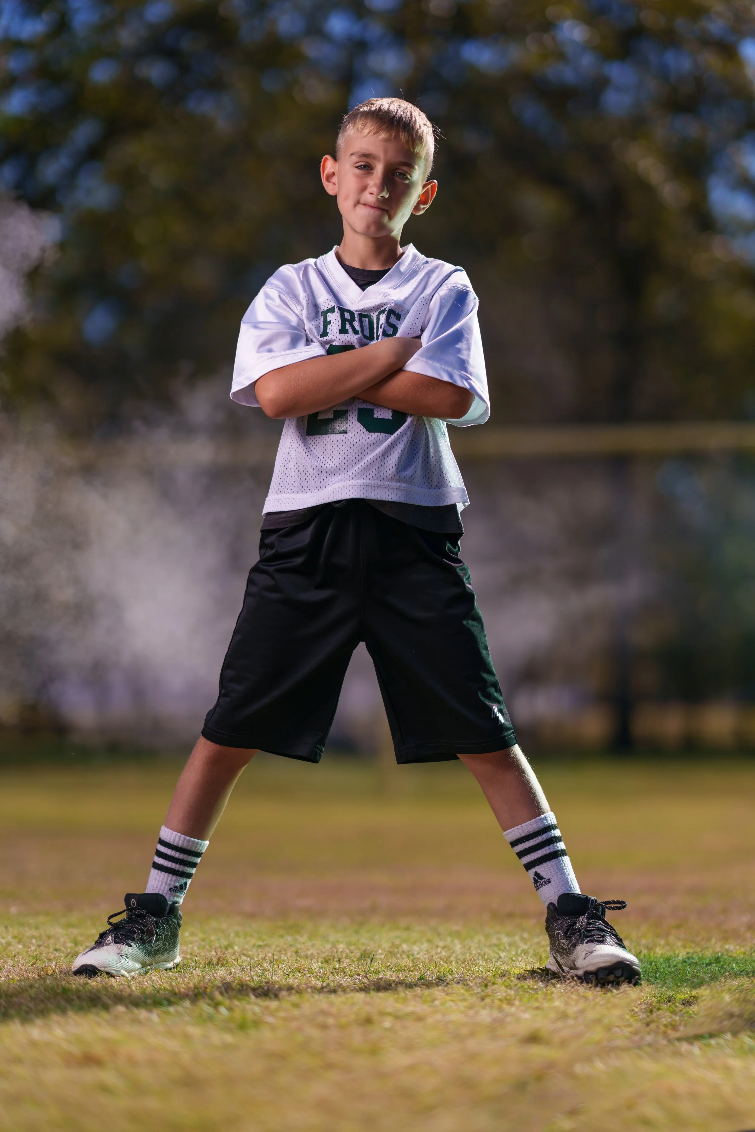 A young boy standing on a grassy field with his arms crossed, wearing a football jersey, shorts, and athletic shoes with socks, outdoors on a sunny day.