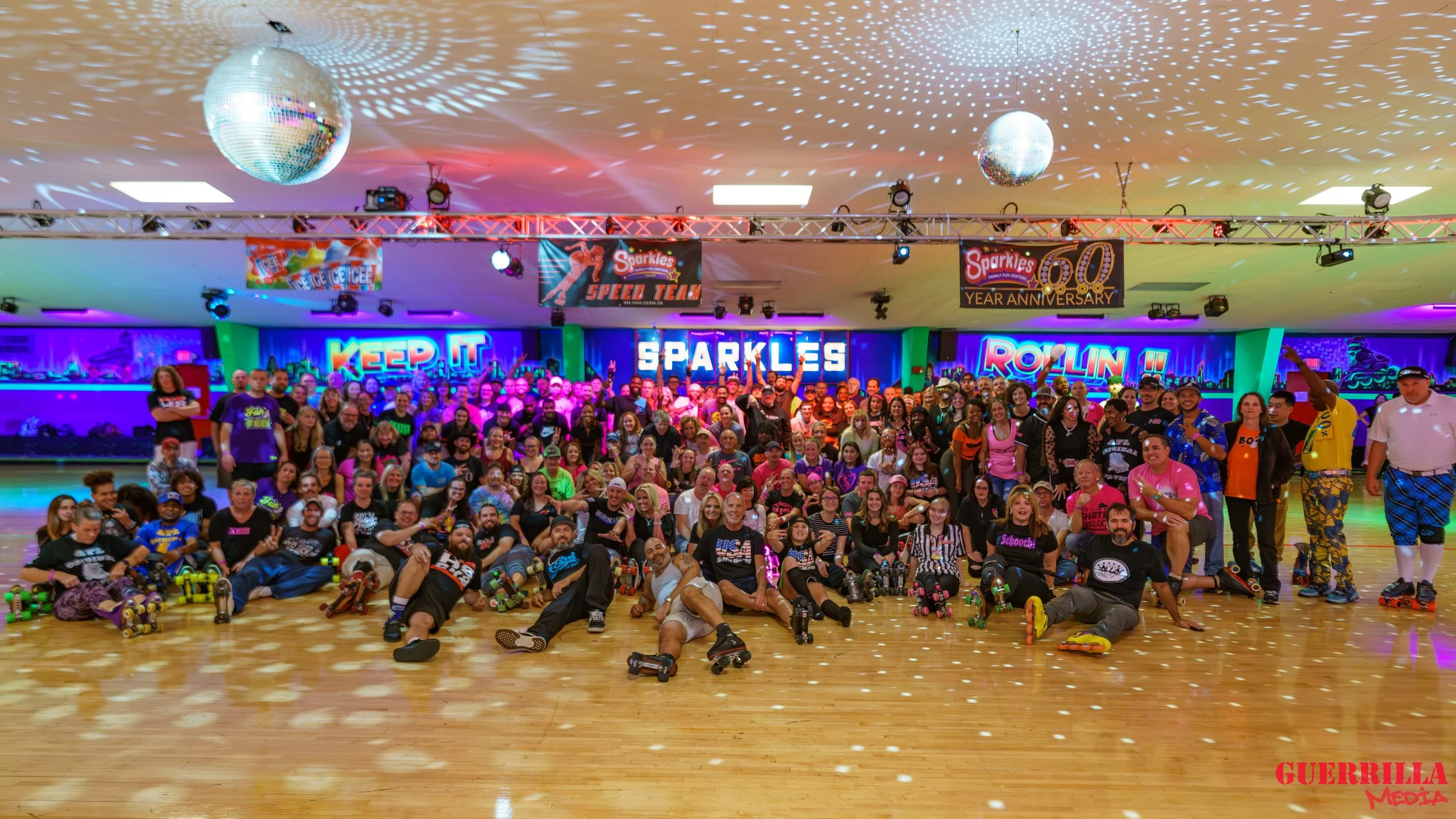 A large group of people gathered in a roller skating rink with neon signs and colorful lighting celebrating an event. The signs read 'Keep It Sparkles,' 'Rollin,' and 'Sparkles 60 Year Anniversary.' Disco balls hang from the ceiling, and the floor is
