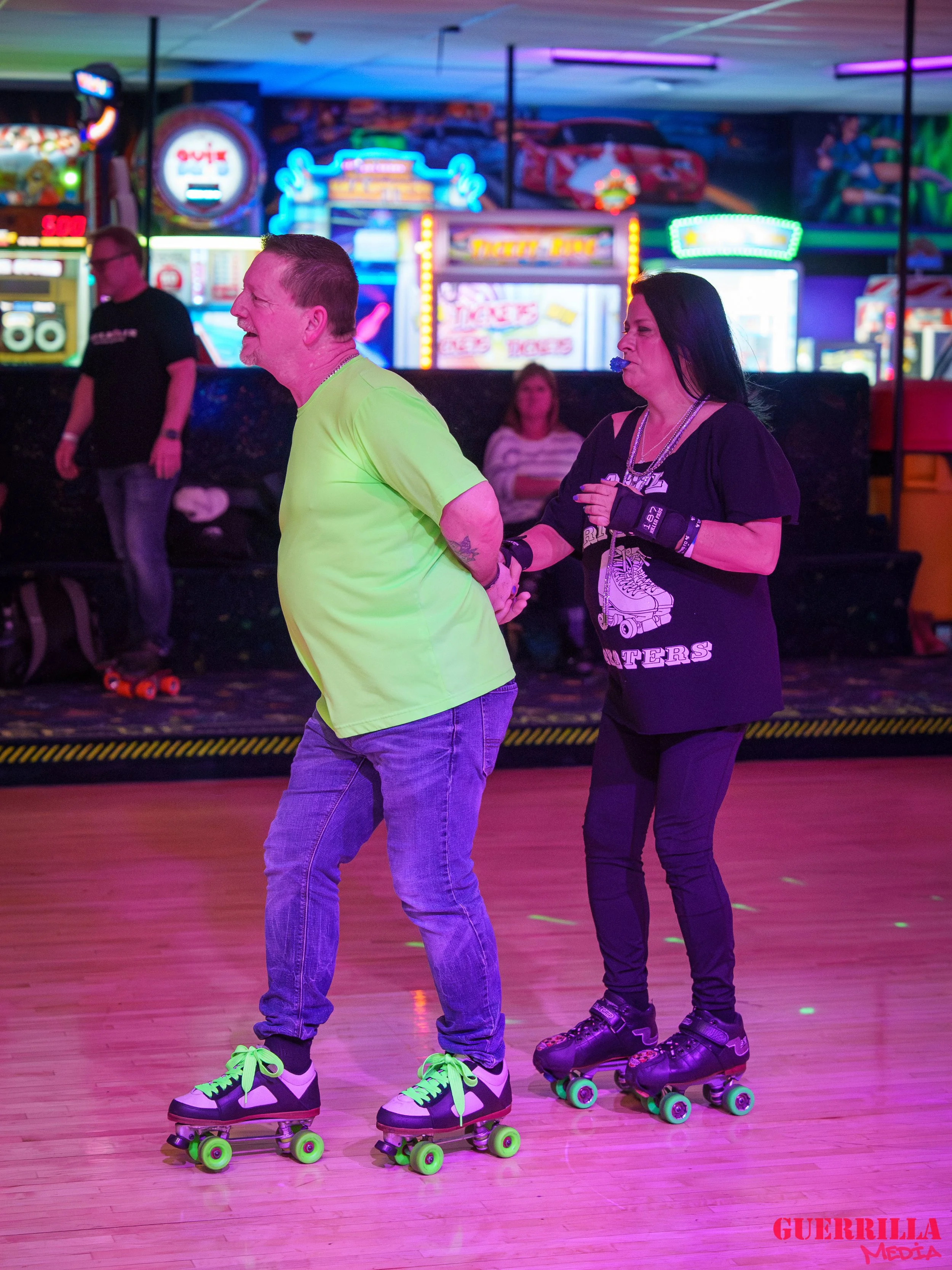 Two people roller skating indoors, holding hands, with neon lights and arcade game signs in the background.