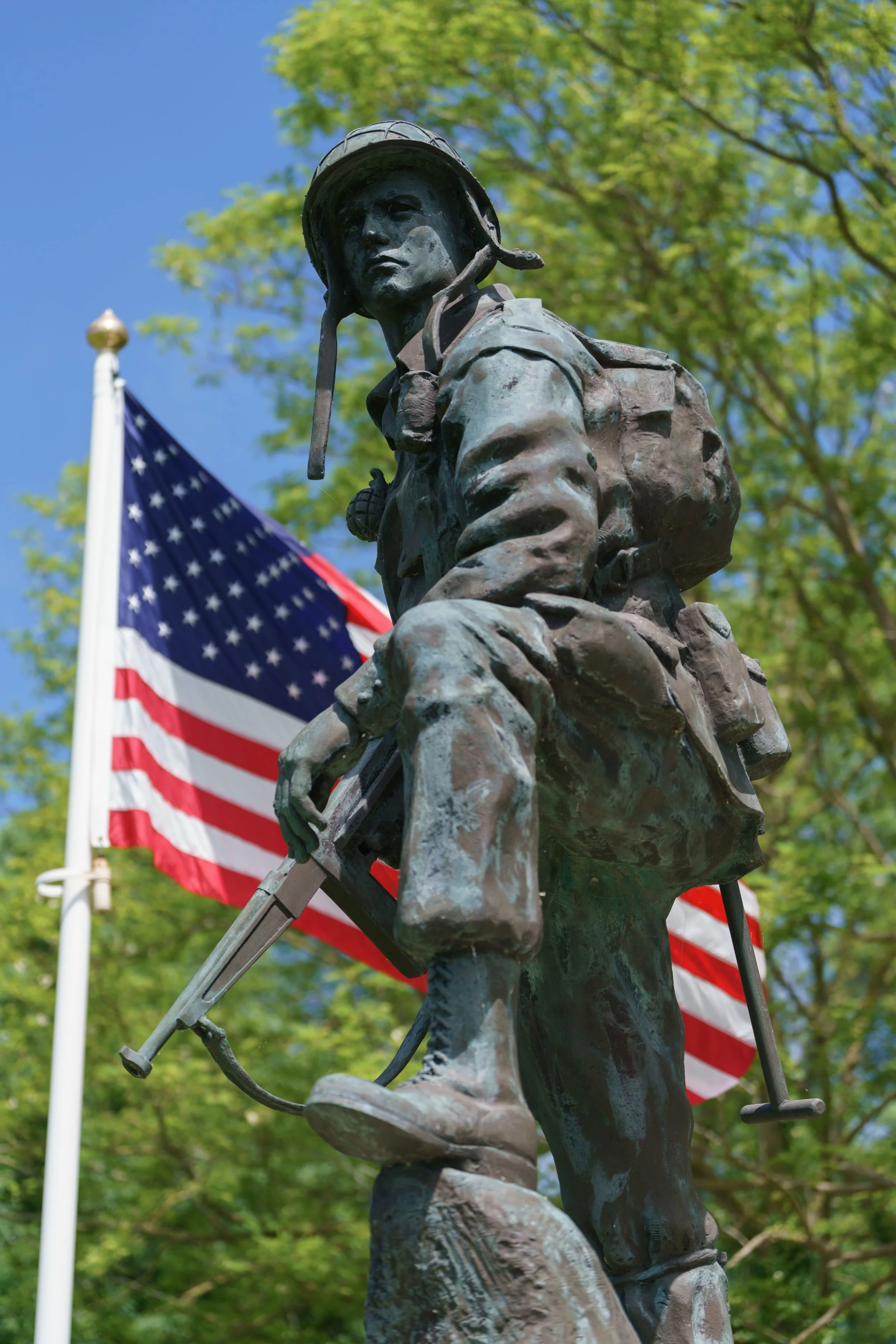 Bronze soldier statue with American flag in the background, green trees, and blue sky