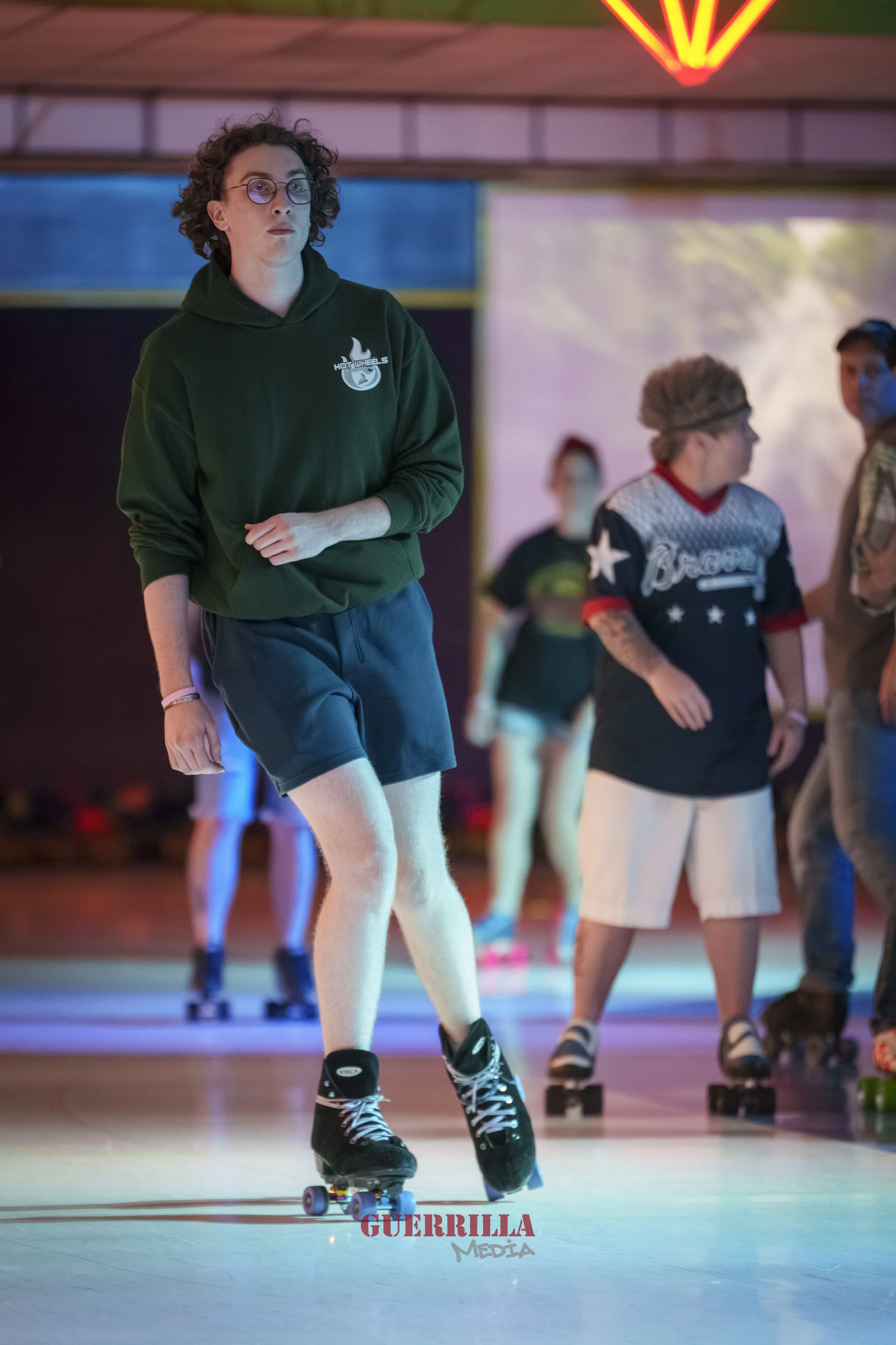 A young man with curly hair, glasses, wearing a green hoodie and shorts, roller skating indoors with a group of people in the background.