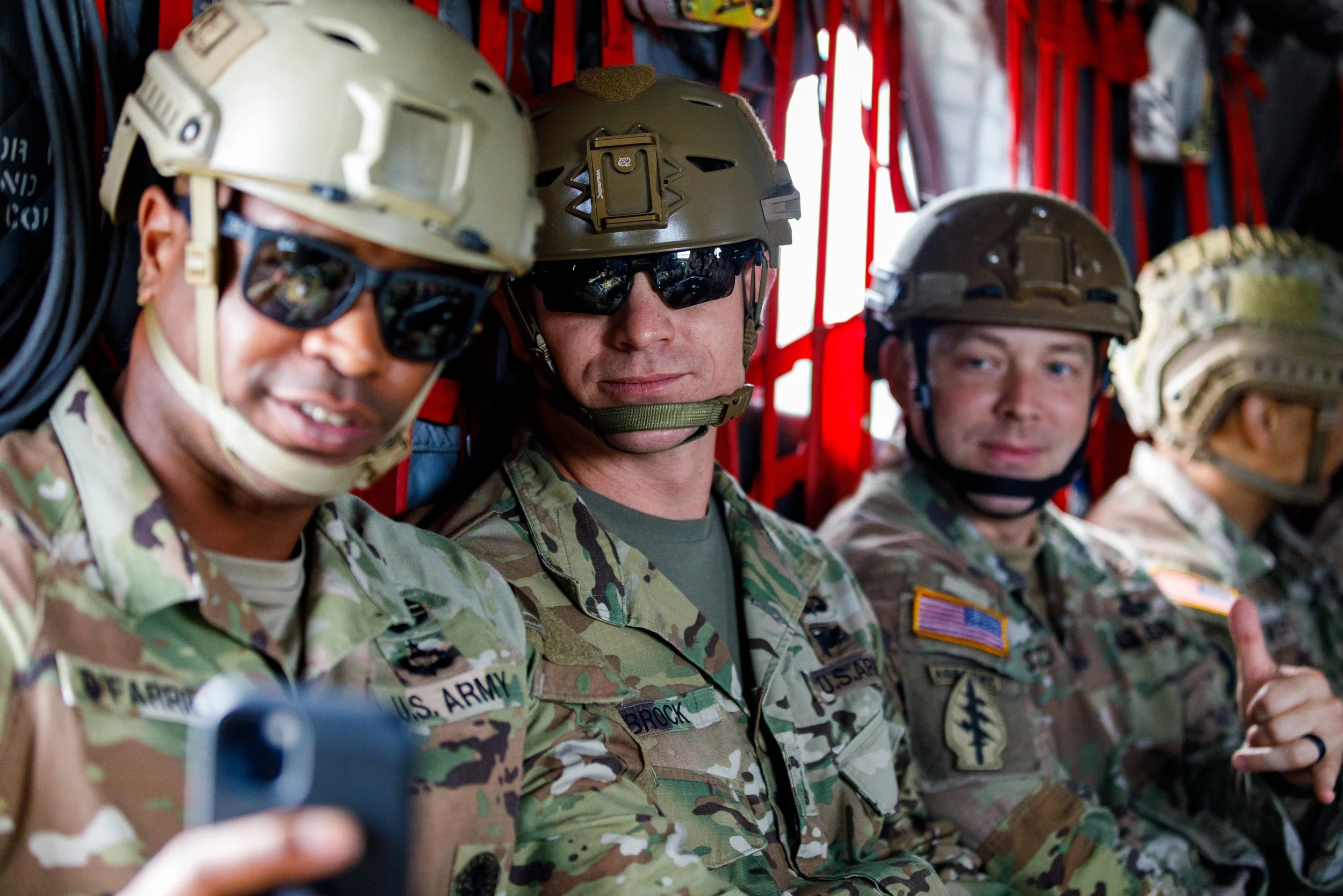 Four soldiers in camouflage uniforms wearing helmets and sunglasses sitting inside a military aircraft.