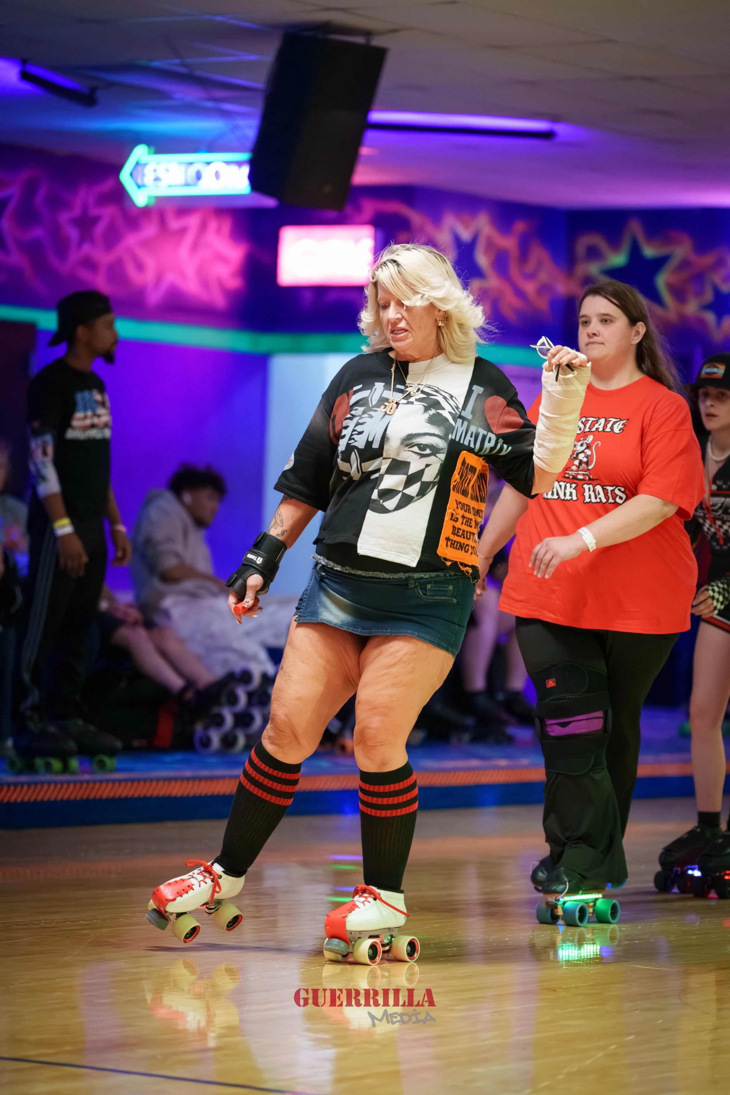 Woman roller skating in a rink with others nearby, neon signs, and colorful graffiti-style wall art in the background.
