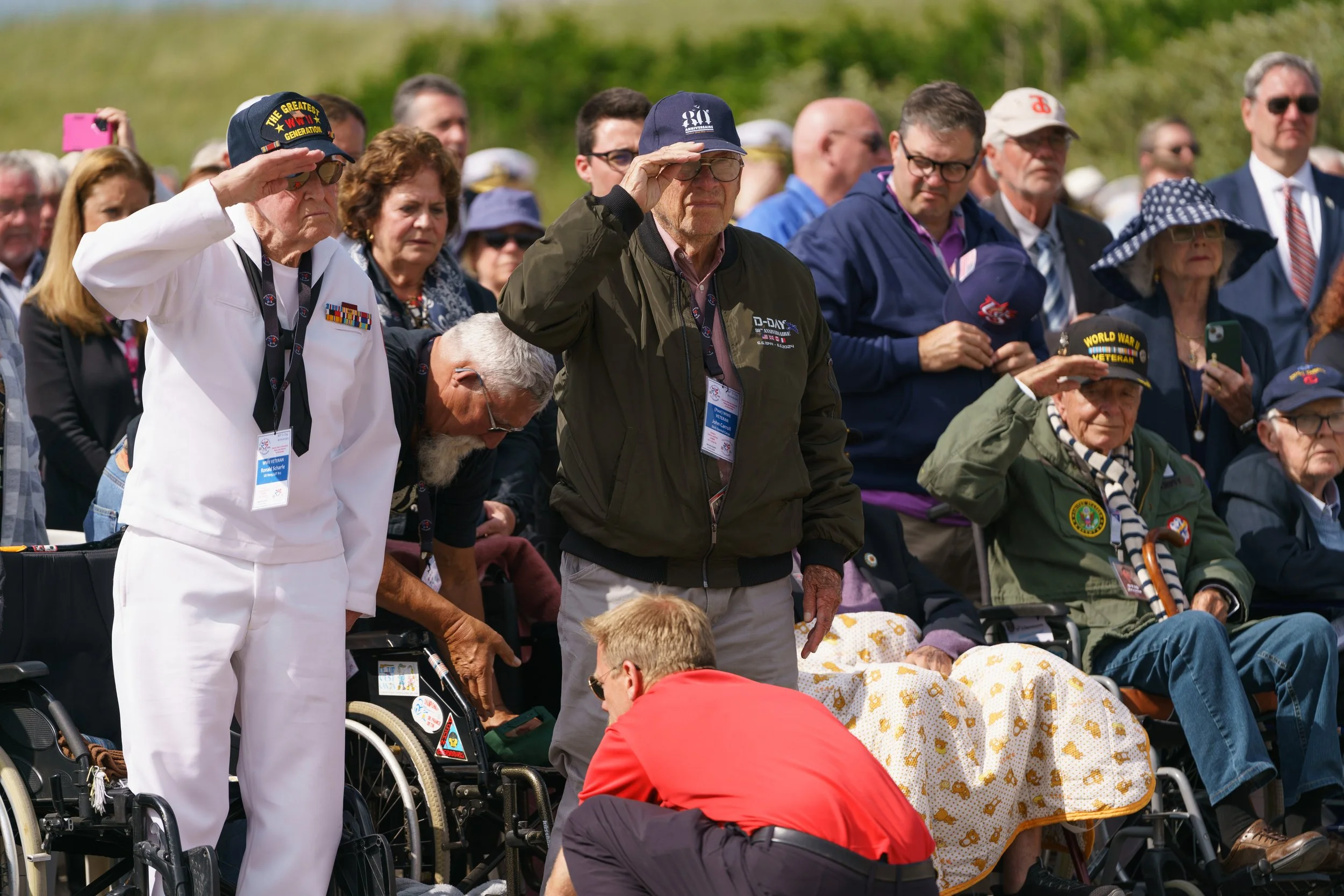 A group of elderly veterans, some in military uniforms and hats, participating in a ceremonial event outdoors. They are saluting and gathering around a seated elderly woman with a blanket, with some holding hats and others taking photos.