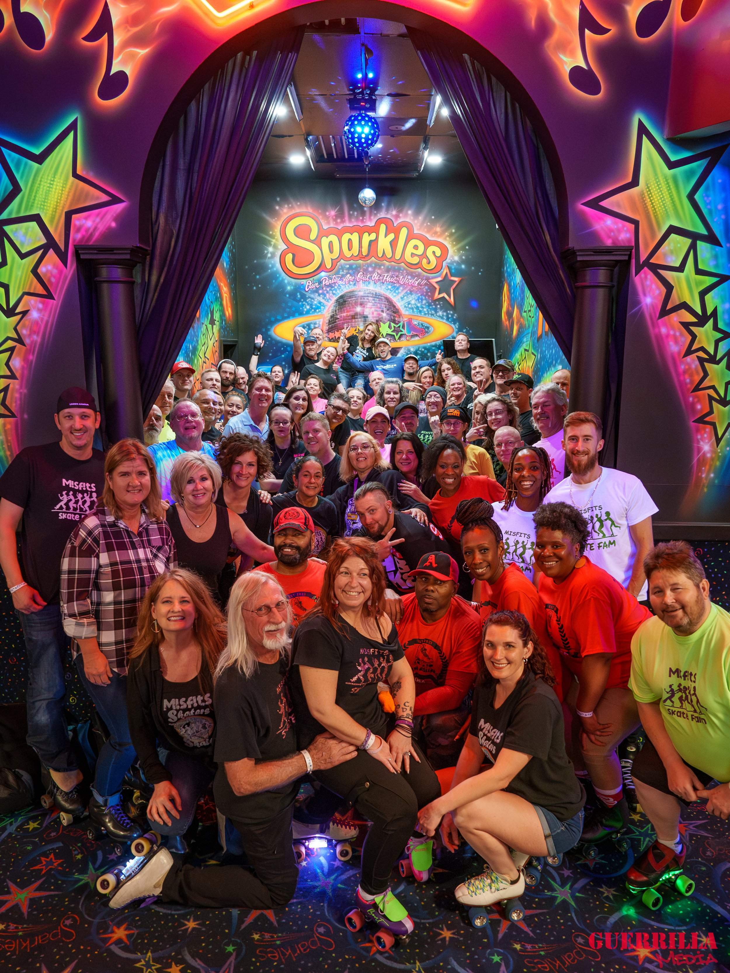 A group of smiling people posing for a photo at a roller skating rink with colorful star and planet themed decorations and a sign that reads 'Sparkles Our Planet Are Out Of This World'.