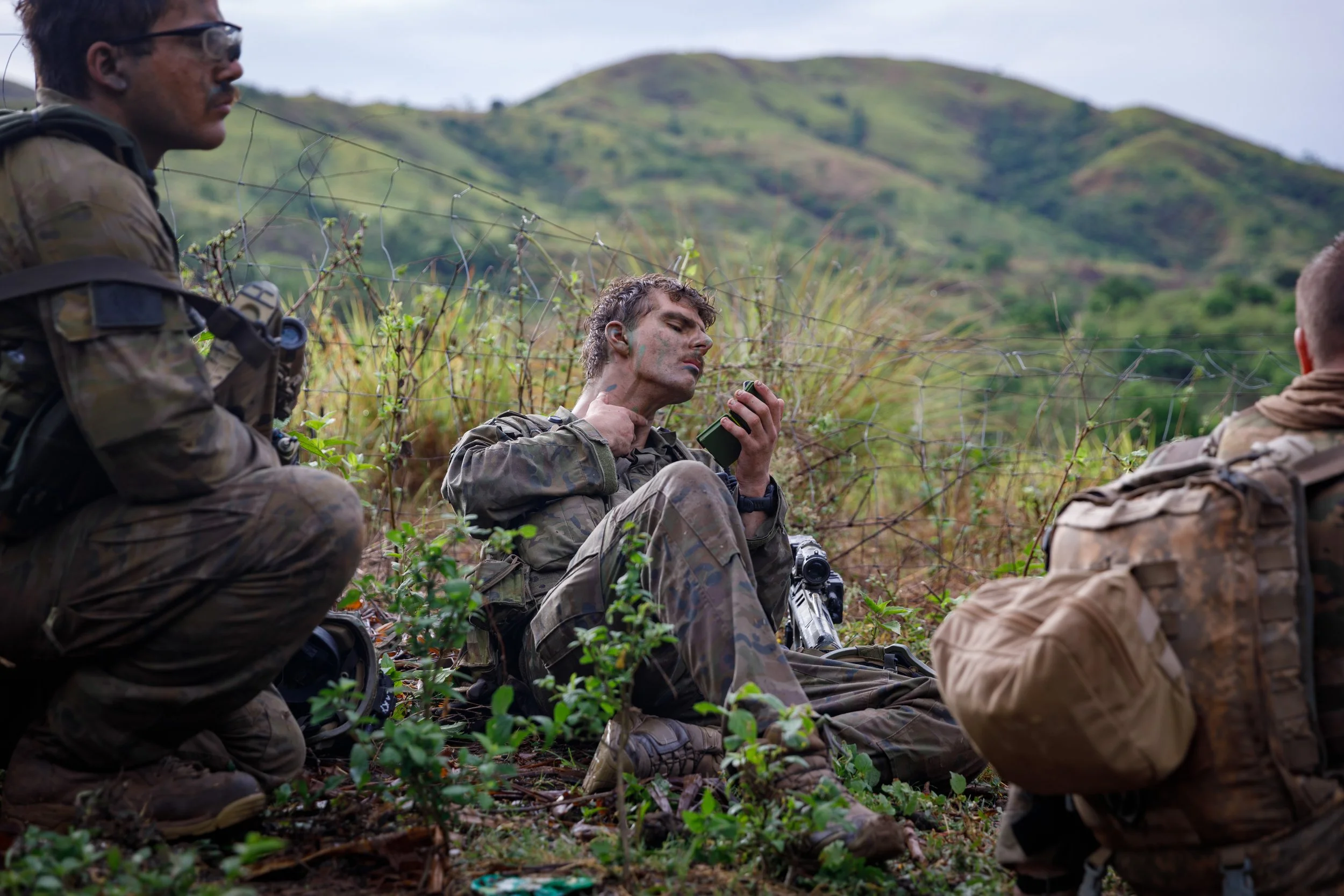 Three soldiers in camouflage gear sit on the ground in a field with green hills in the background; one looks at a device, another is crouching, and the third is sitting with his back to the camera.