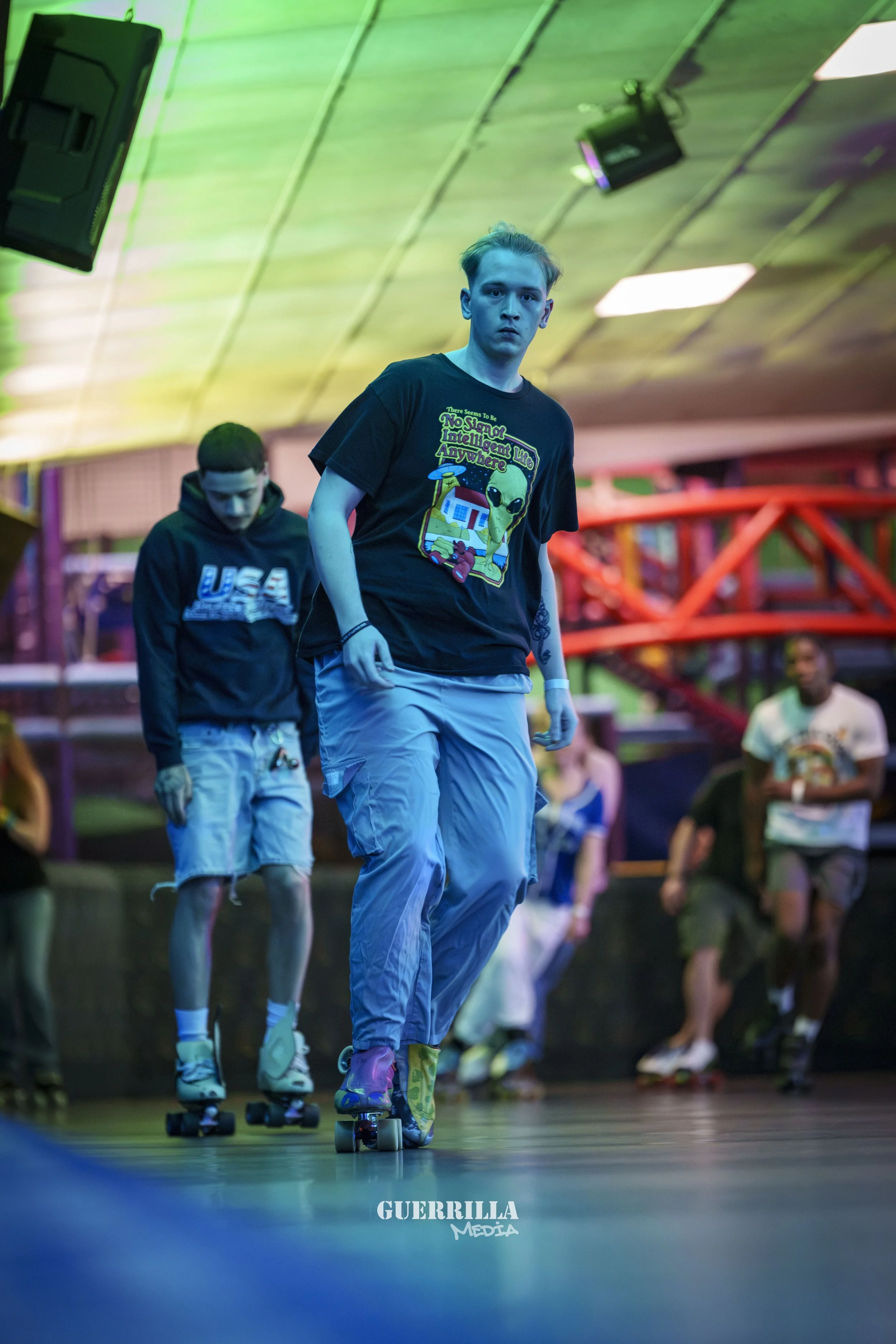 Two young men roller skating indoors with colorful lighting and a red bridge structure in the background.