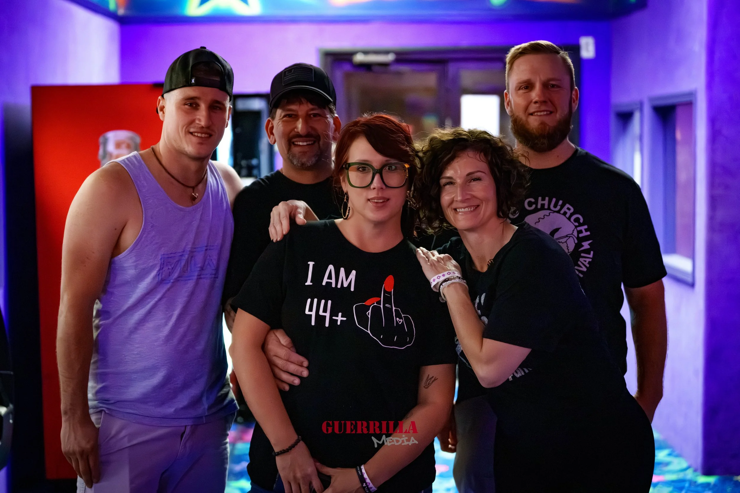 Group of five people at a social event, with purple lighting, smiling and posing for the photo, one person wearing an 'I am 44+' t-shirt.