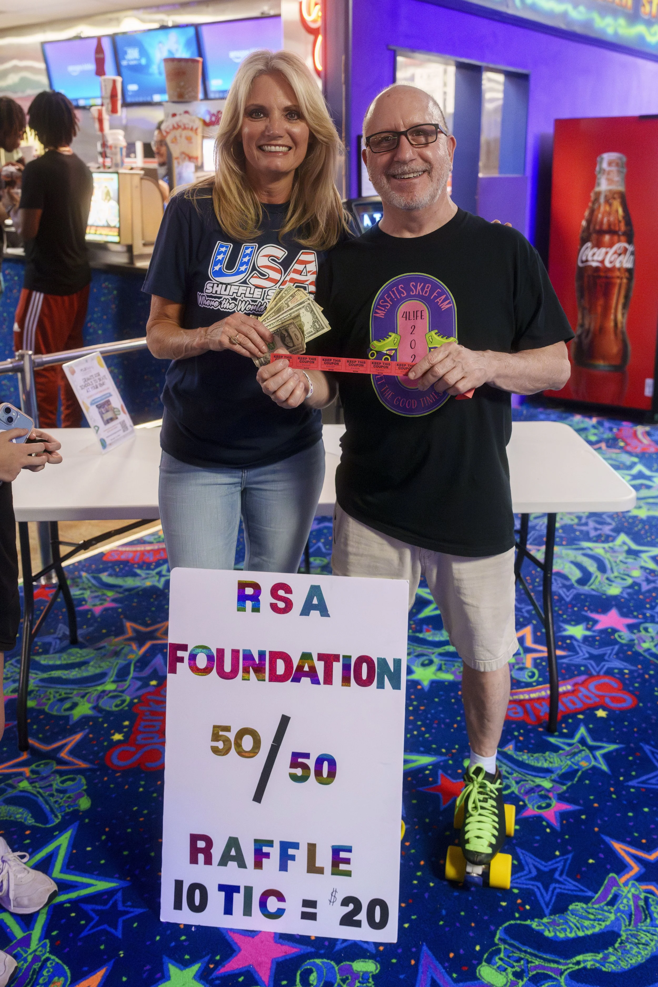 A smiling woman and man are standing together at a raffle event inside an arcade or entertainment venue. The woman is holding a stack of cash and raffle tickets, while the man is holding tickets and standing on roller skates. There is a colorful sign