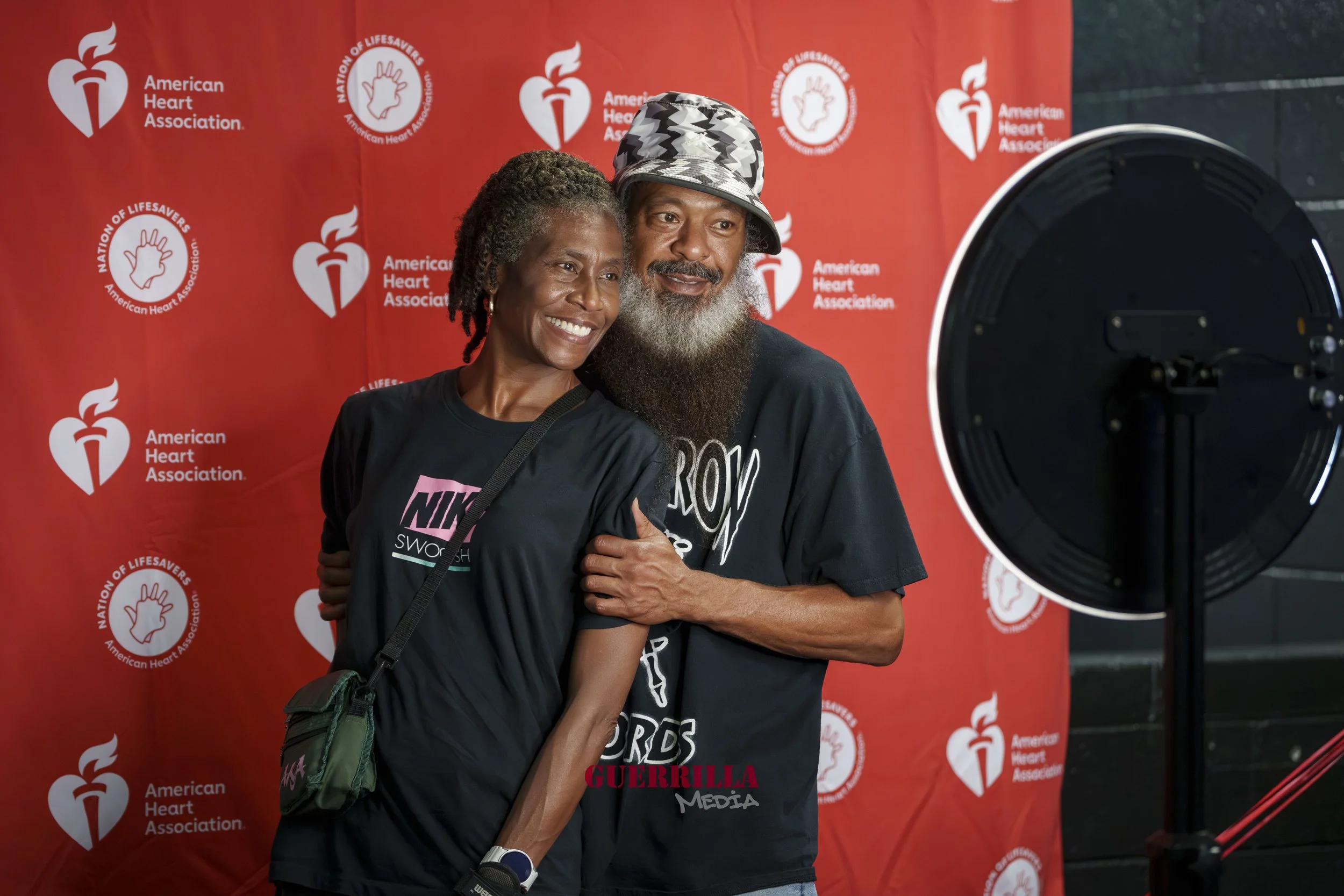 A smiling woman and a bearded man taking a selfie in front of a red backdrop with the American Heart Association logo. The woman is wearing a black Nike t-shirt and the man is wearing a black t-shirt and a patterned hat.