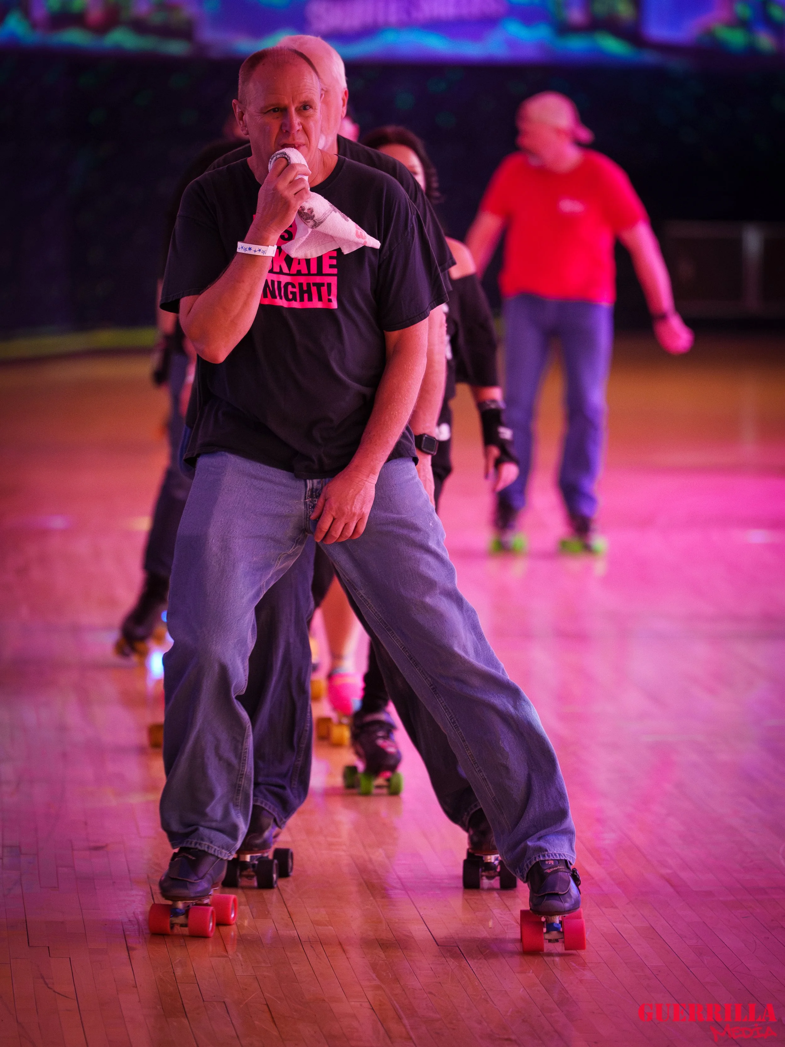 People roller skating indoors on a polished wooden floor with colorful lighting, including a man in the foreground drinking from a water bottle with a towel.