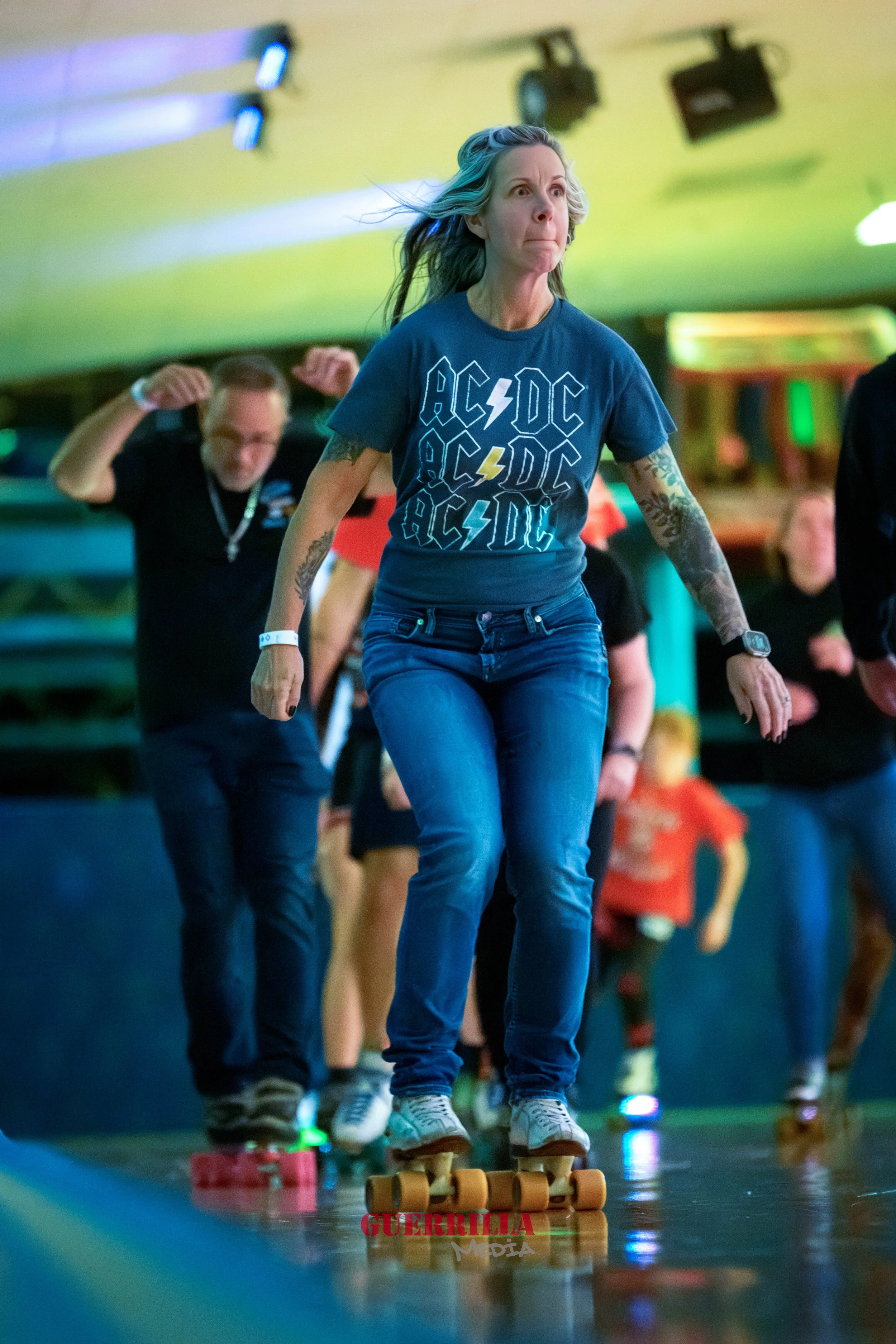 A woman skating on roller skates at an indoor roller skating rink, wearing a blue AC/DC t-shirt. She is surrounded by other skaters in the background.