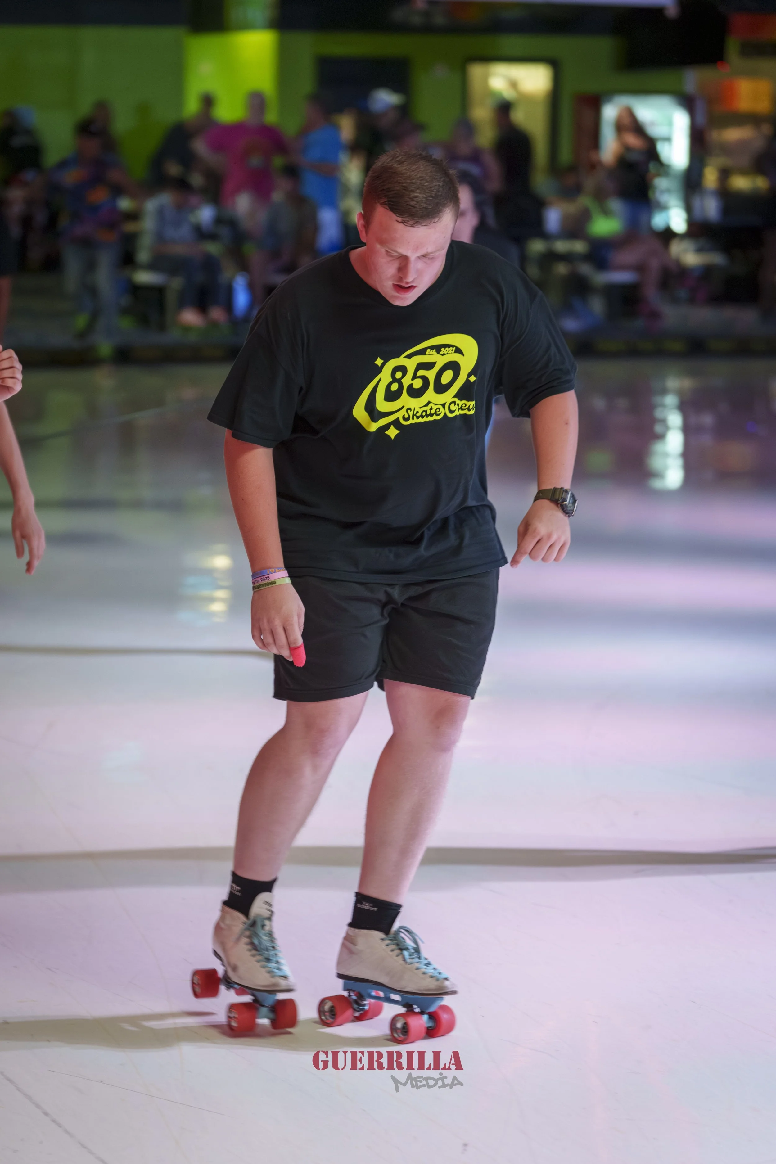 A person roller skating indoors, wearing a black t-shirt with '850 Skate Crew' printed on it, black shorts, and beige sneakers with blue laces. There are people and bright green walls in the background.