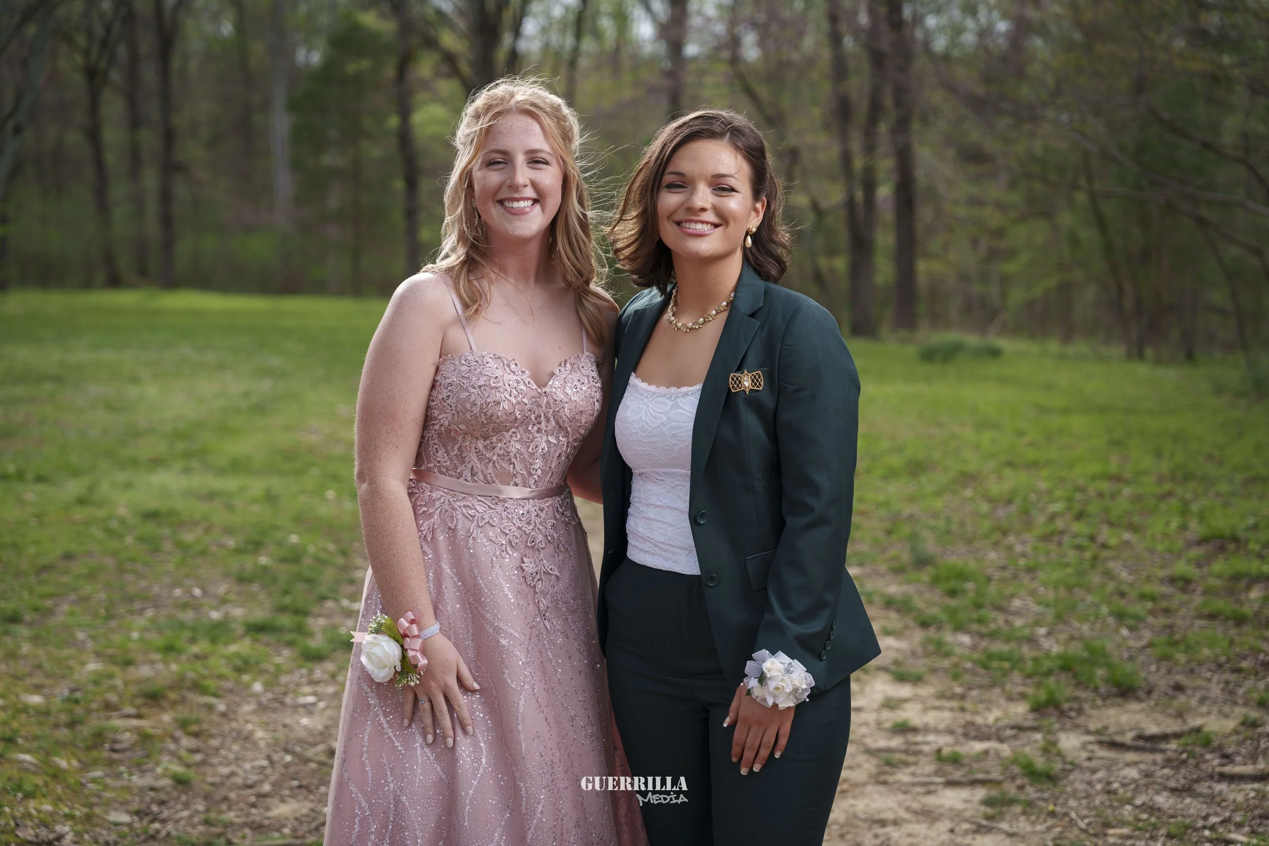 Two young women smiling and standing outdoors in a wooded area, dressed in formal attire. One wears a pink lace gown with a corsage on her wrist, the other wears a dark green blazer with a brooch and a white top, both with floral wrist corsages.