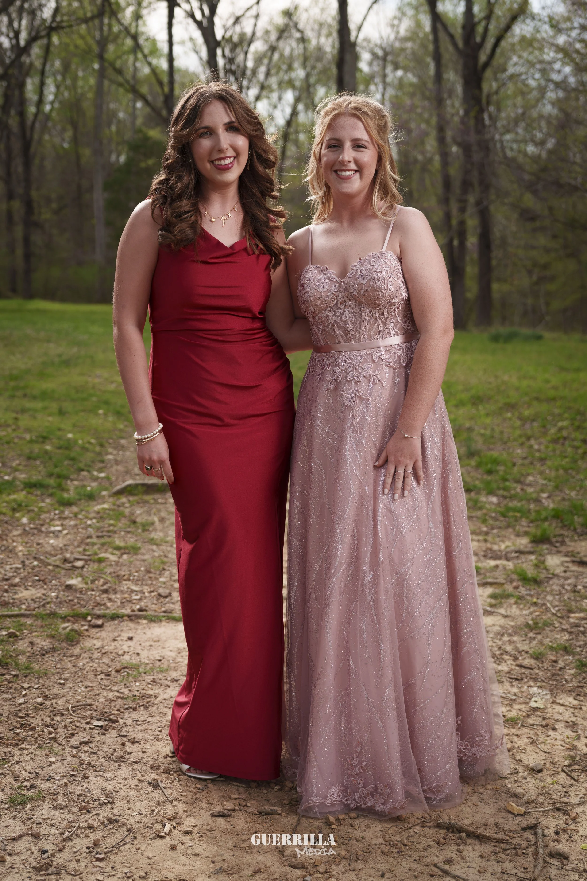 Two women in formal dresses standing outdoors on a dirt path, surrounded by trees and greenery, smiling at the camera.