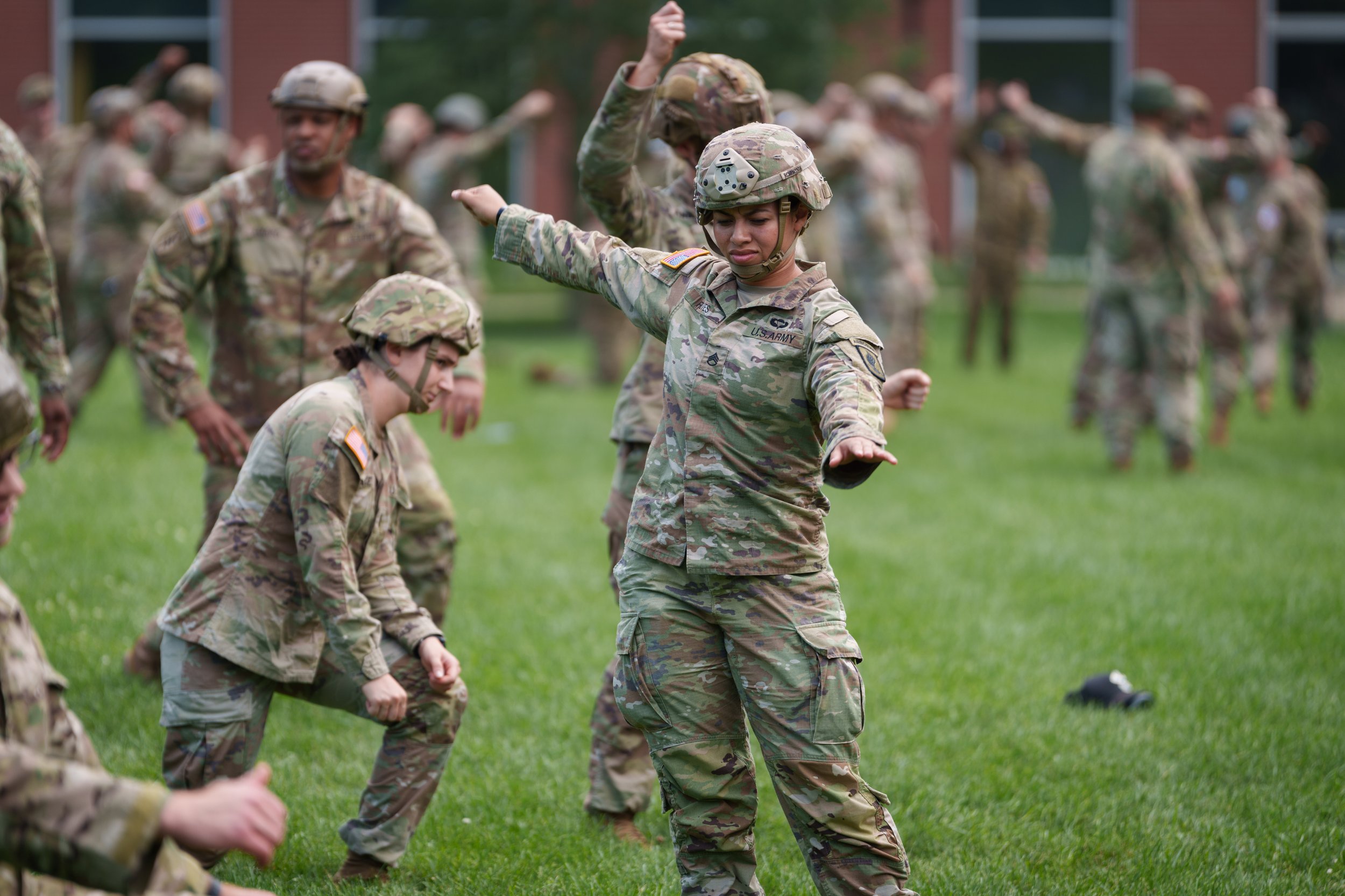 U.S. Army soldiers in camouflage field uniforms practicing martial arts or self-defense techniques on a grassy field, with one soldier demonstrating a stance.