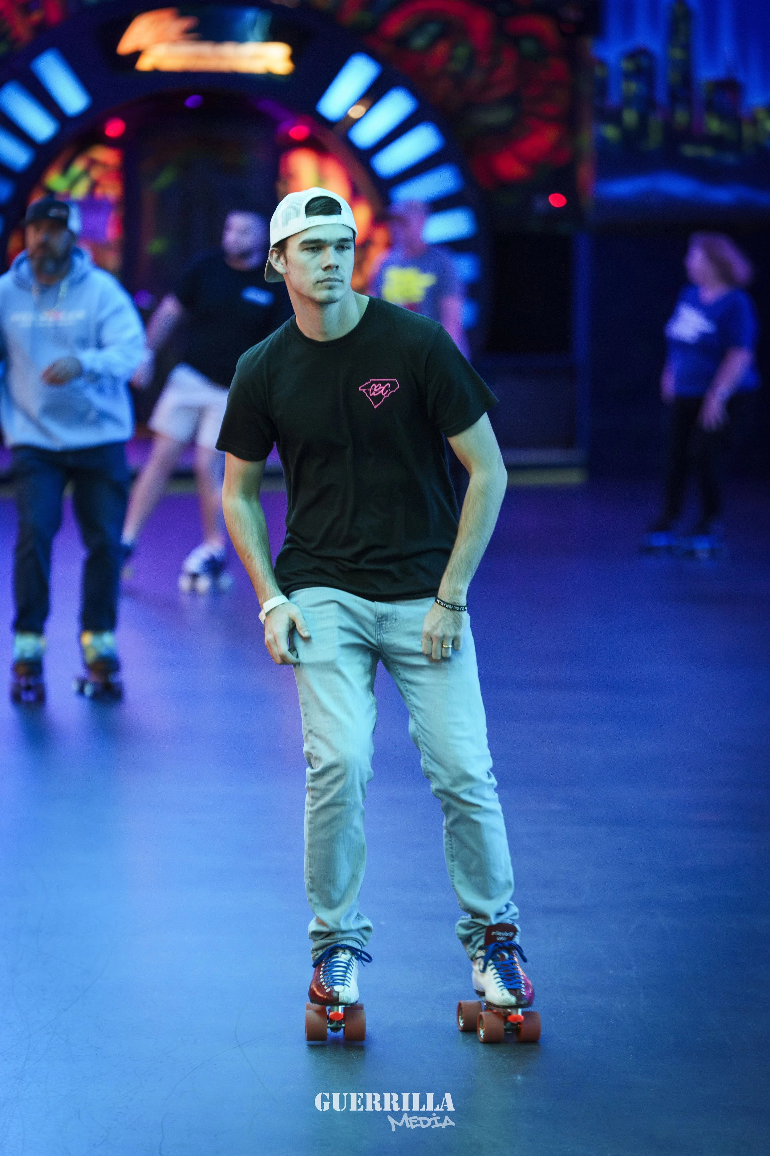 A young man roller skating on a roller rink with colorful neon lights, wearing a black T-shirt, light jeans, and a white cap backwards.