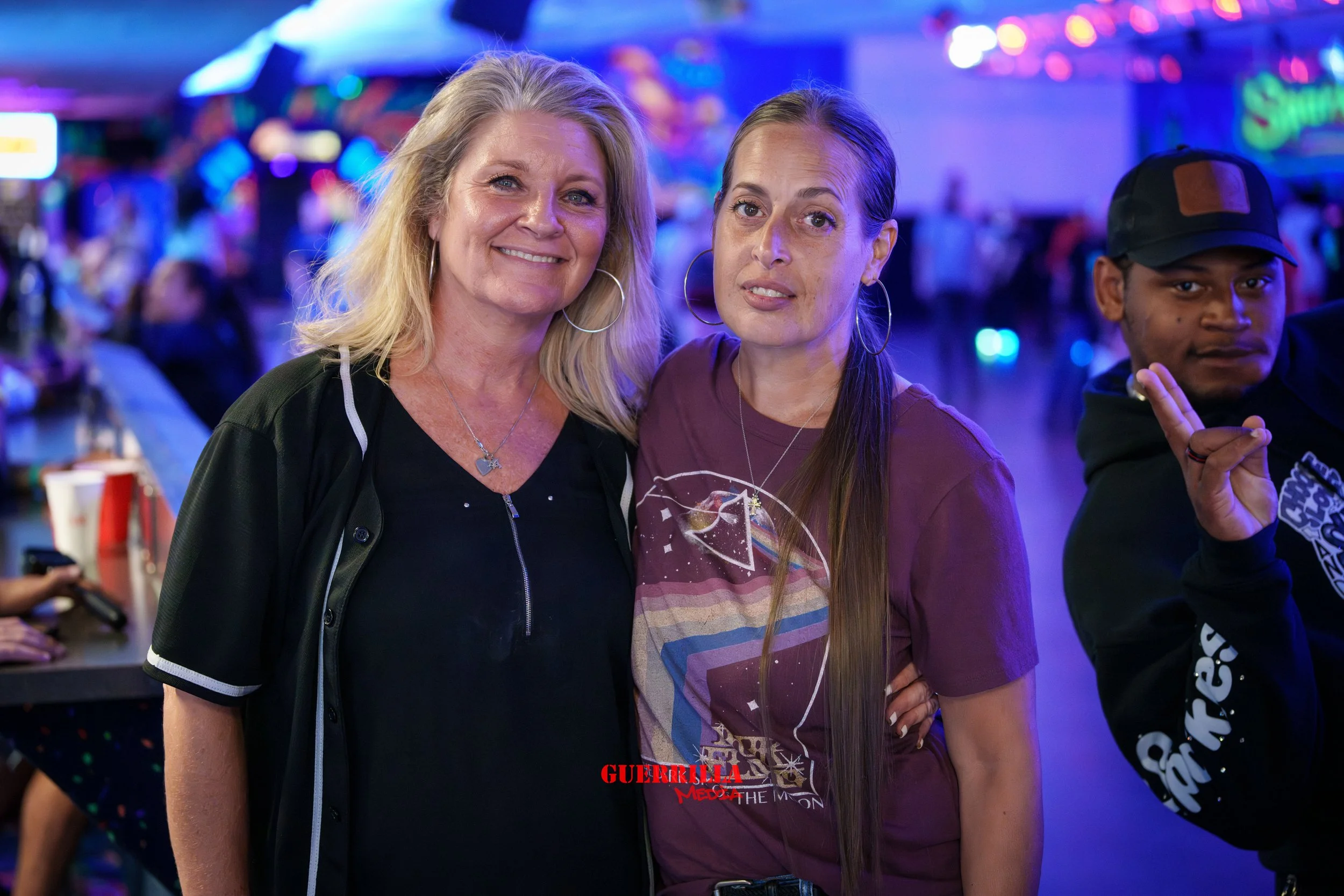 Two women and a young man in a neon-lit arcade. The women are smiling, one with long blonde hair and the other with long brown hair. The young man is making a peace sign. Bright colorful signs and arcade games are visible in the background.